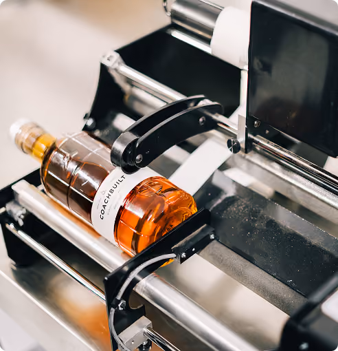 Glass bottle of amber liquid labeled 'Coach & Built' being labeled by an automated machine.