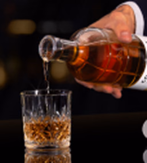 A hand pouring whiskey from a glass bottle into a crystal tumbler glass.