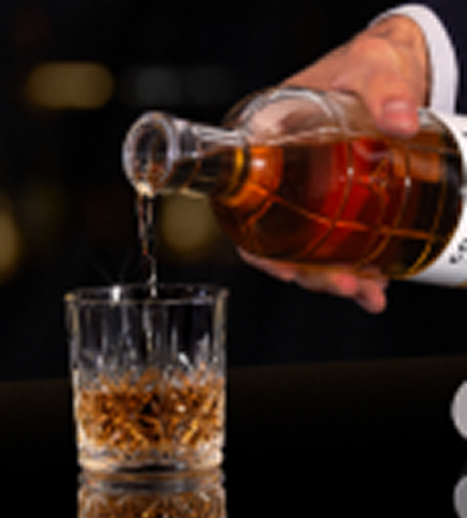 A hand pouring whiskey from a glass bottle into a crystal tumbler glass.