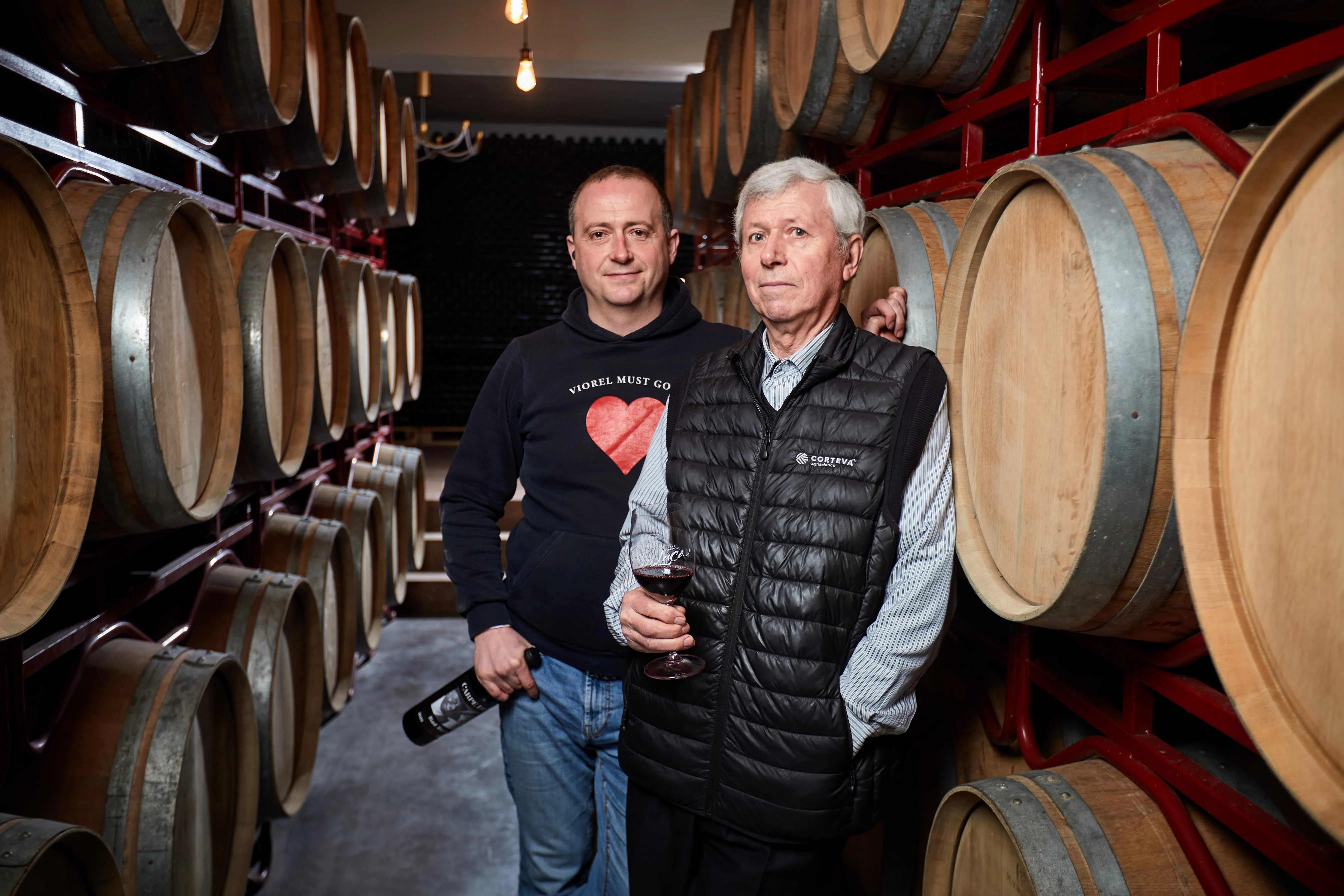 Ion Luca Jr. and Ion Luca Sr. in a barrel cellar, one holding a glass of red wine and the other a bottle