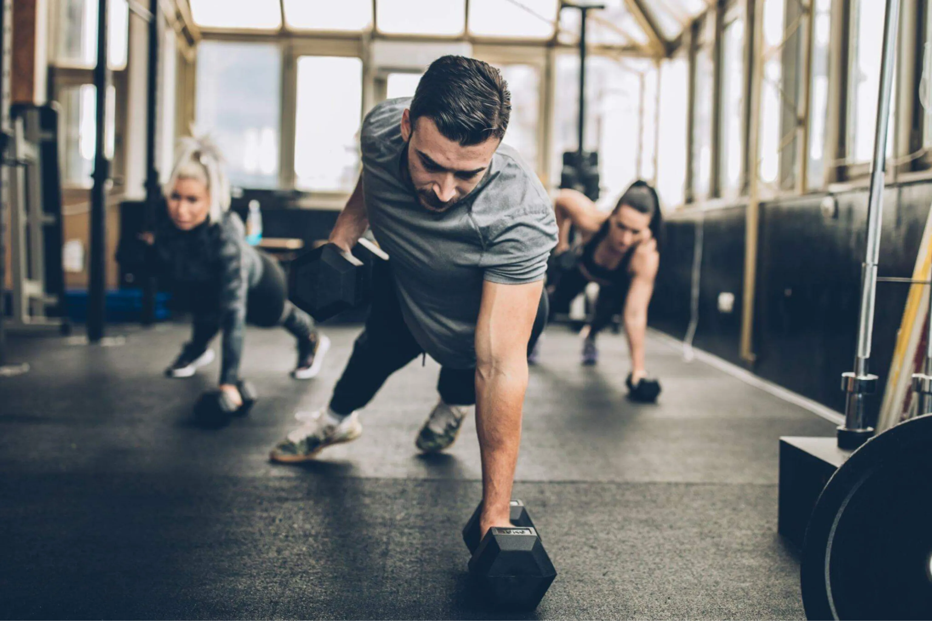 Three people exercising indoors, performing a plank row with dumbbells on a gym floor, with sunlight coming through large windows in the background.