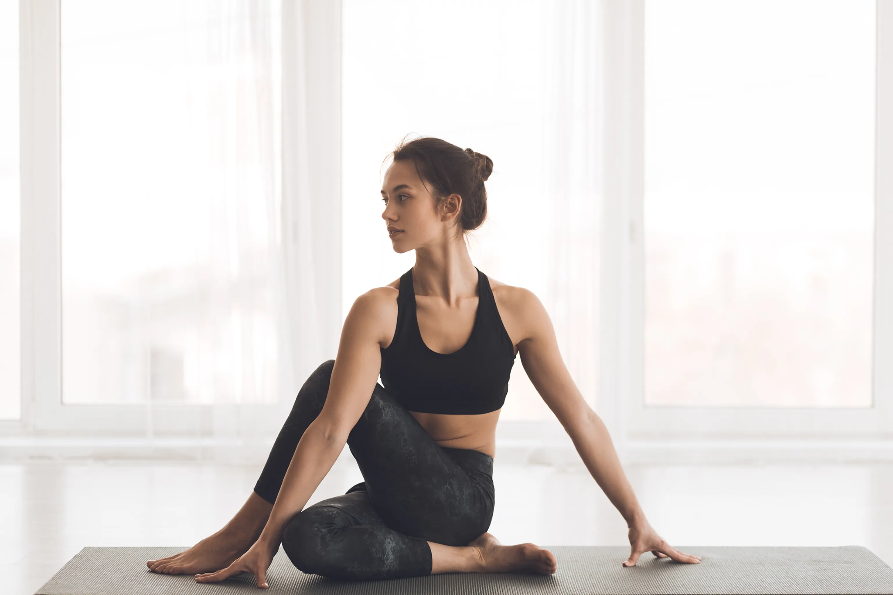Fit woman in black workout clothes sits in a seated yoga twist on a mat by bright windows, conveying calm, balance, and focused wellness.