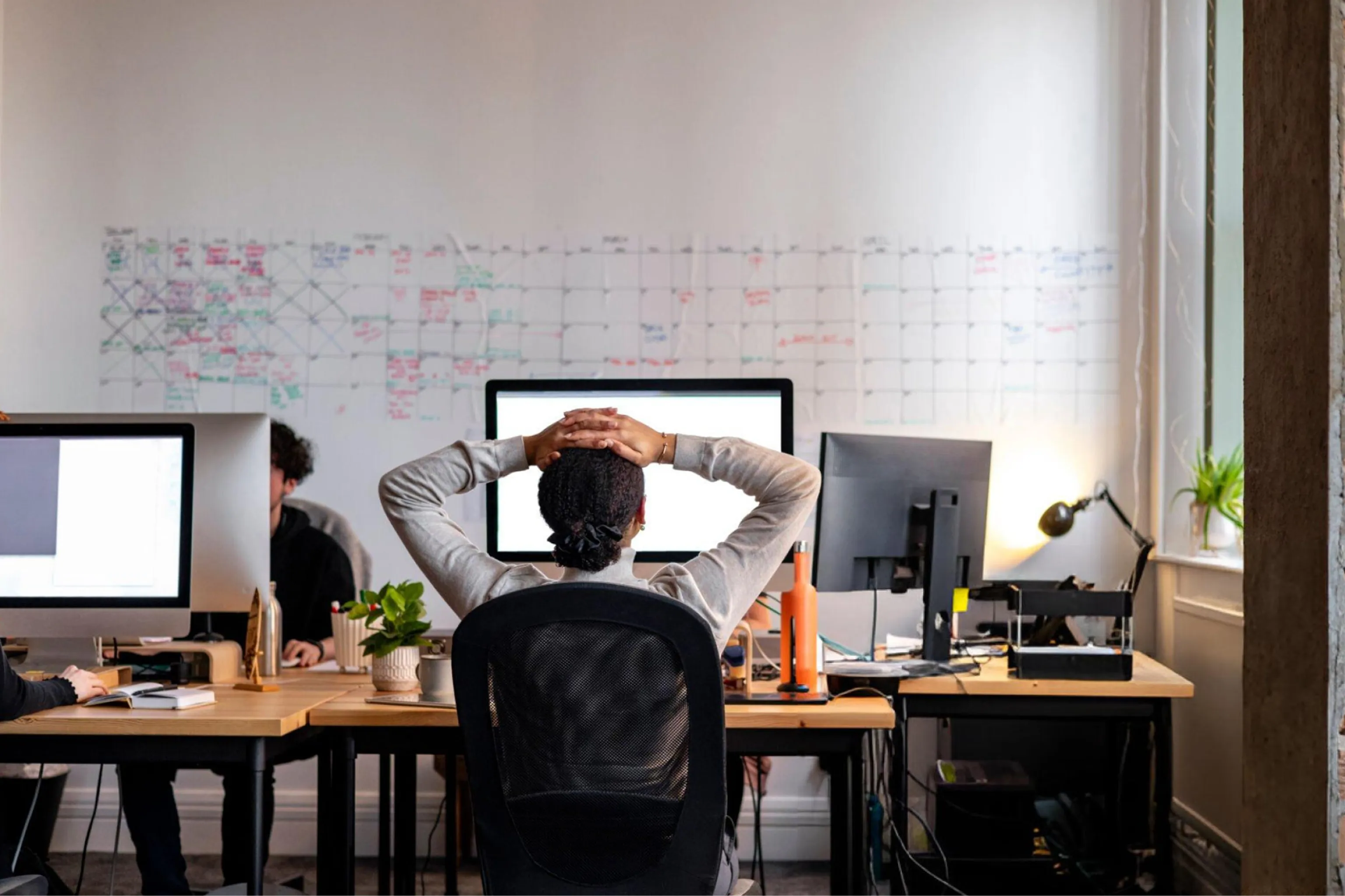 Open-plan office with a relaxed employee leaning back, hands behind their head, surrounded by monitors, coworkers, plants, and a planning wall.