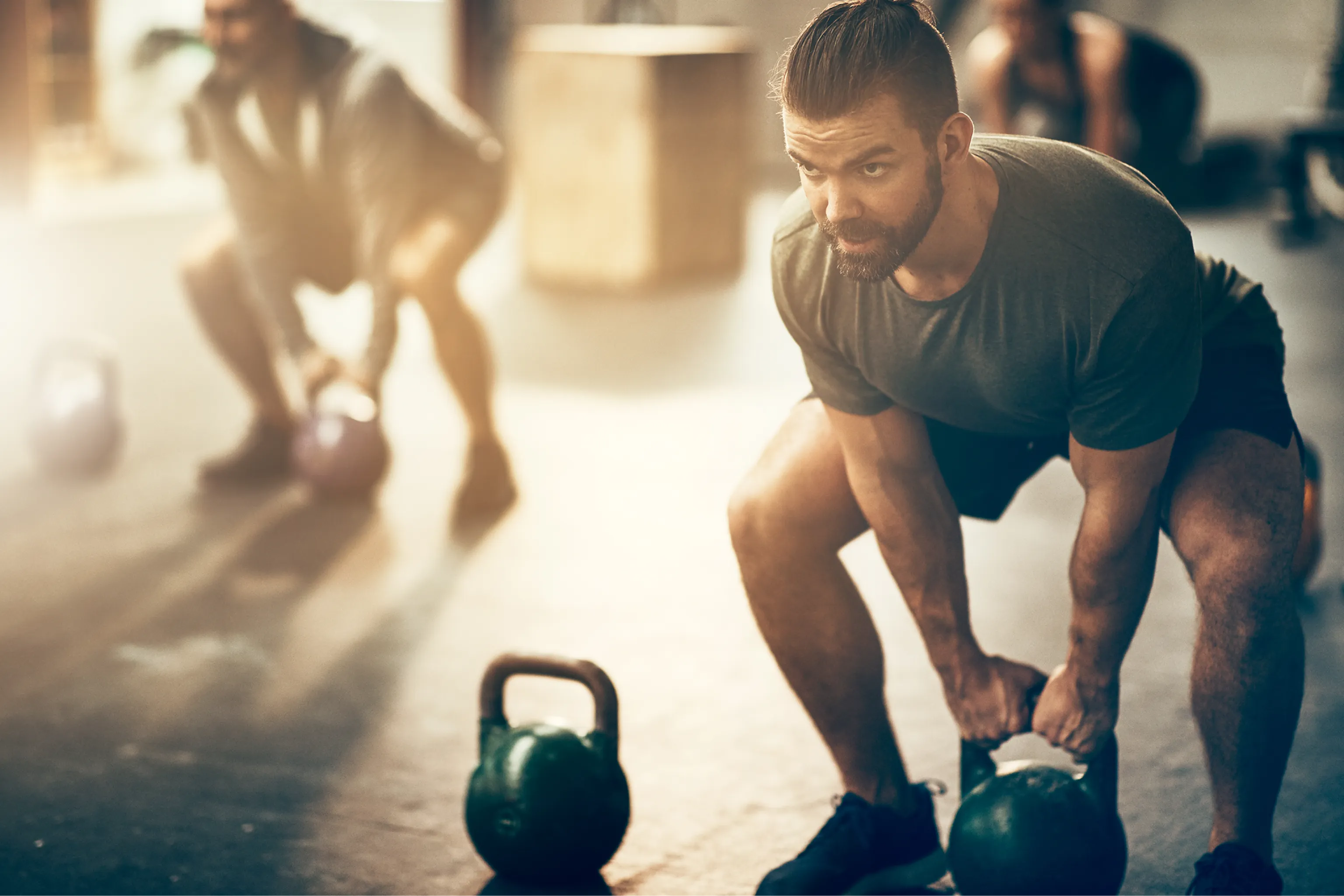 Bearded man lifting a kettlebell during an intense gym workout, with another athlete exercising in the sunlit background.