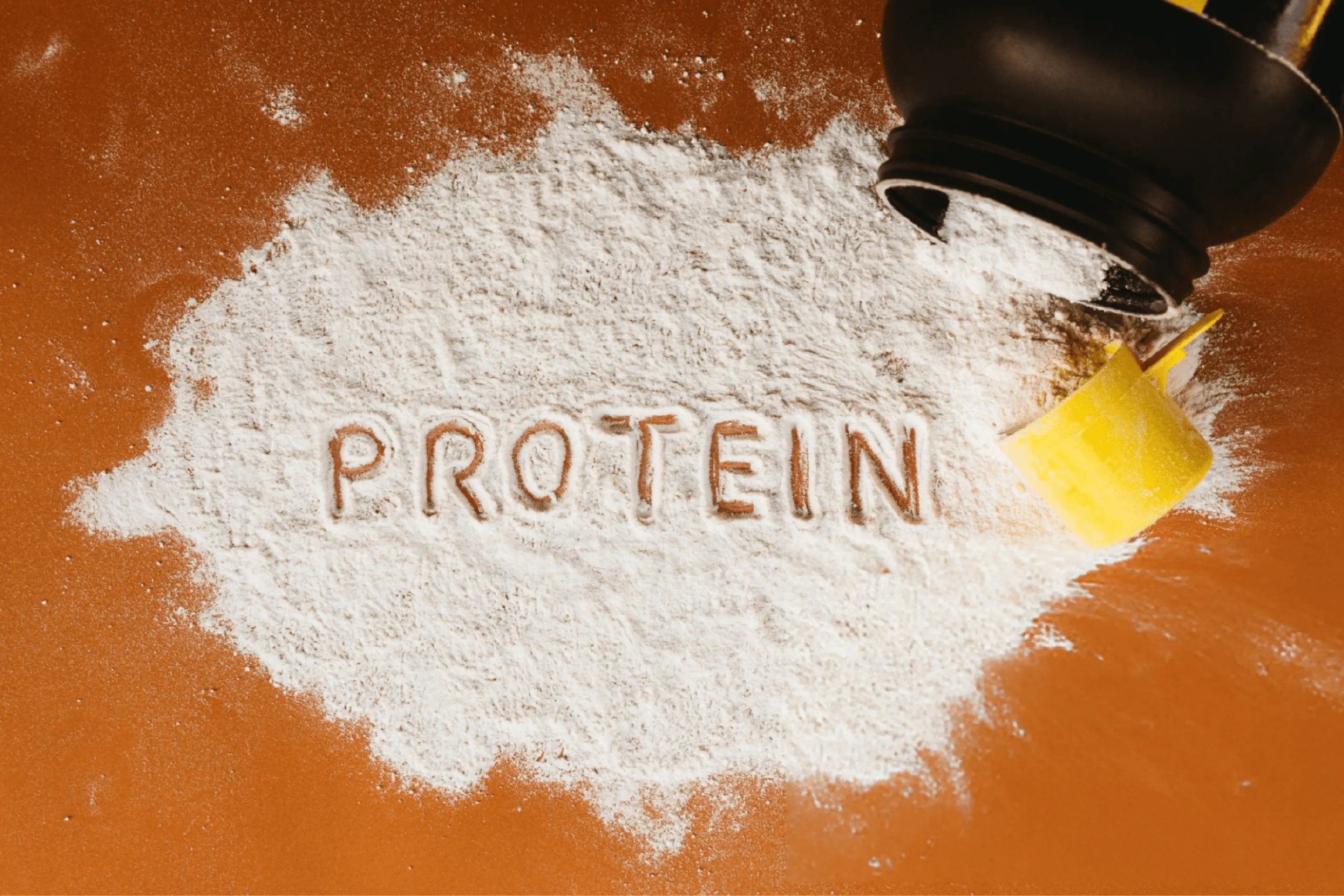 White protein powder forms a mound on a brown surface, with “PROTEIN” traced in it and a black-and-yellow scoop spilling powder.
