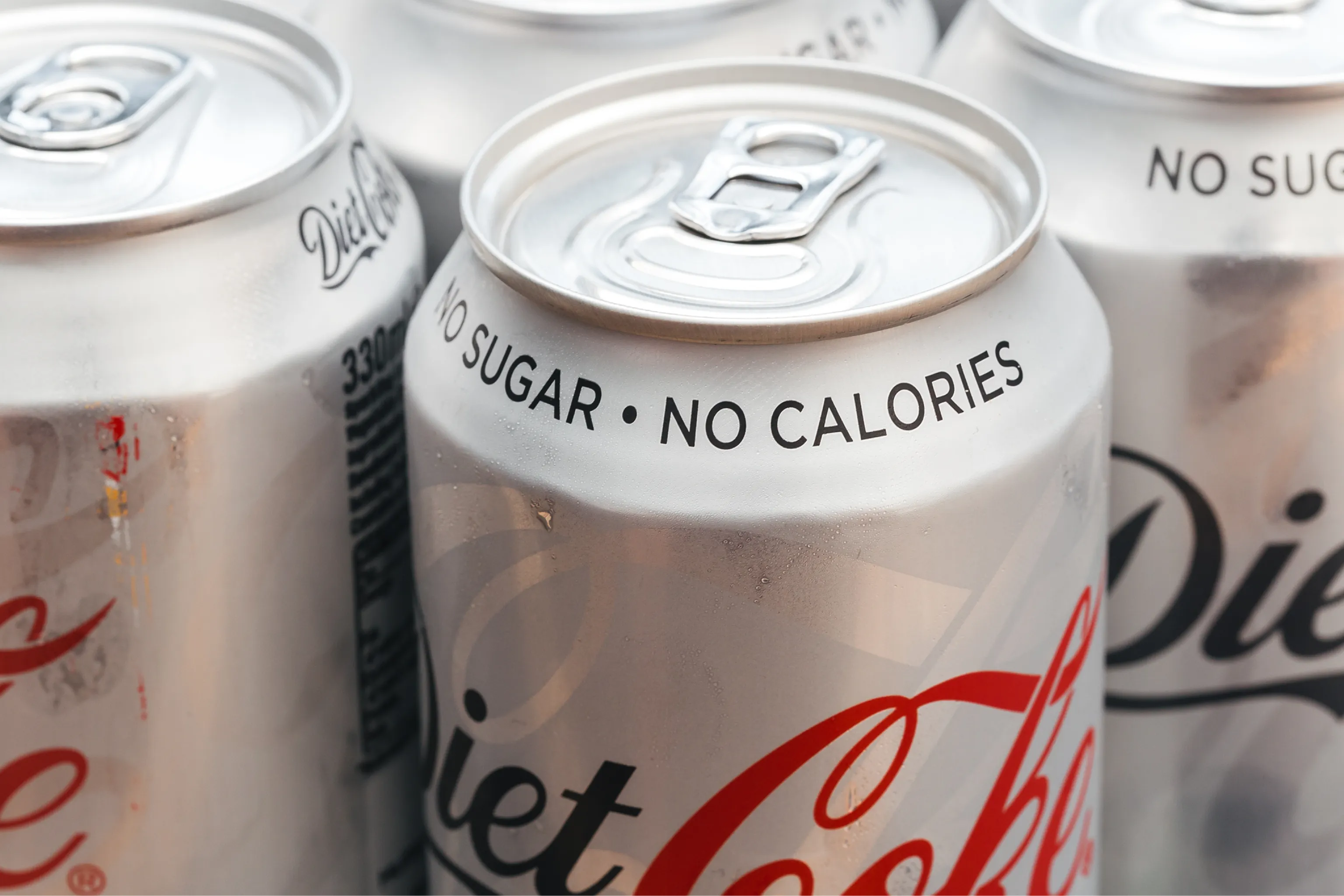 Close-up of silver Diet Coke cans, featuring “NO SUGAR • NO CALORIES” labels and pull tabs, conveying a crisp, refreshing tone.