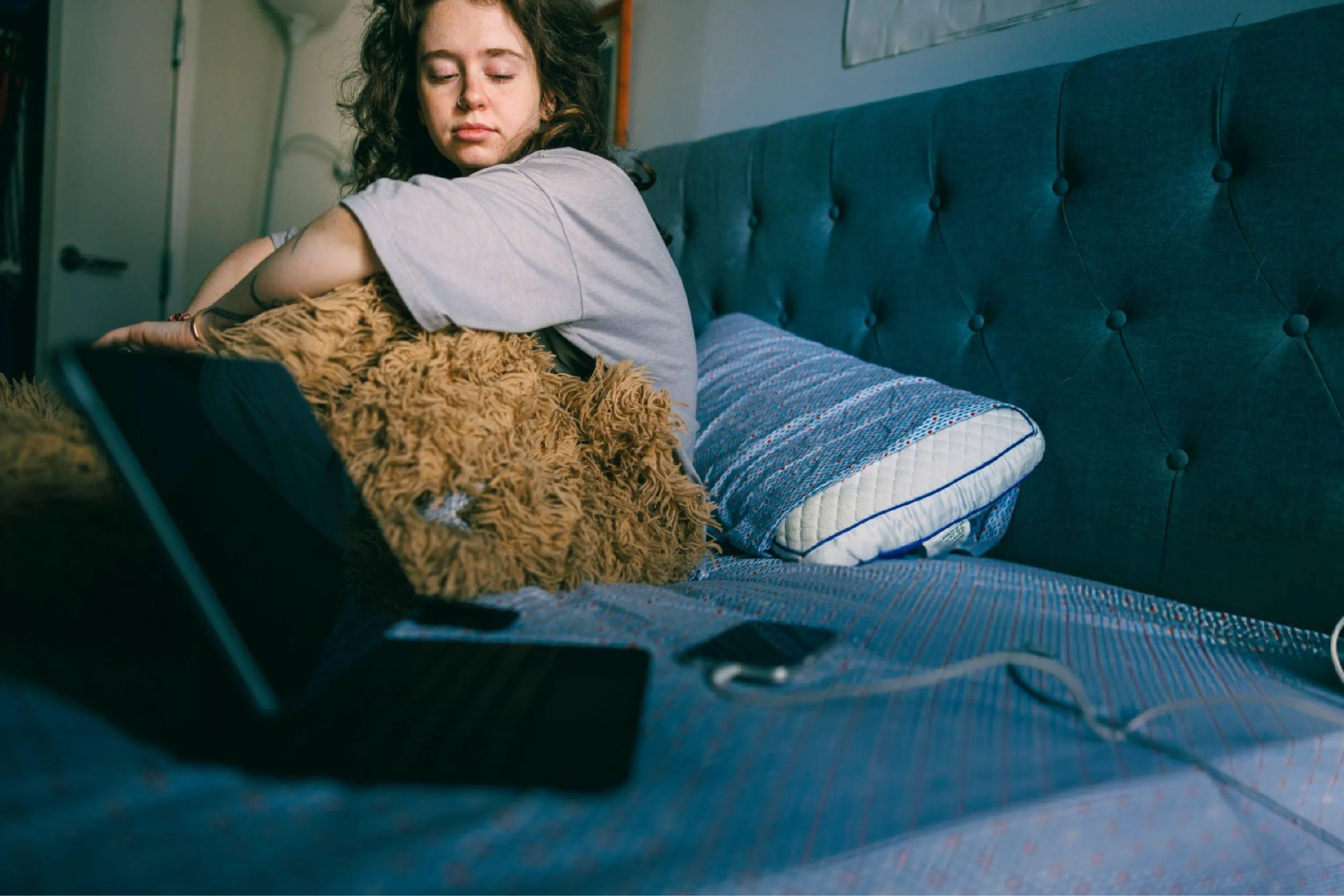 Sleepy young woman rests on a bed beside an open laptop, hugging a shaggy pillow in a dim bedroom; soft light creates a calm, tired mood.