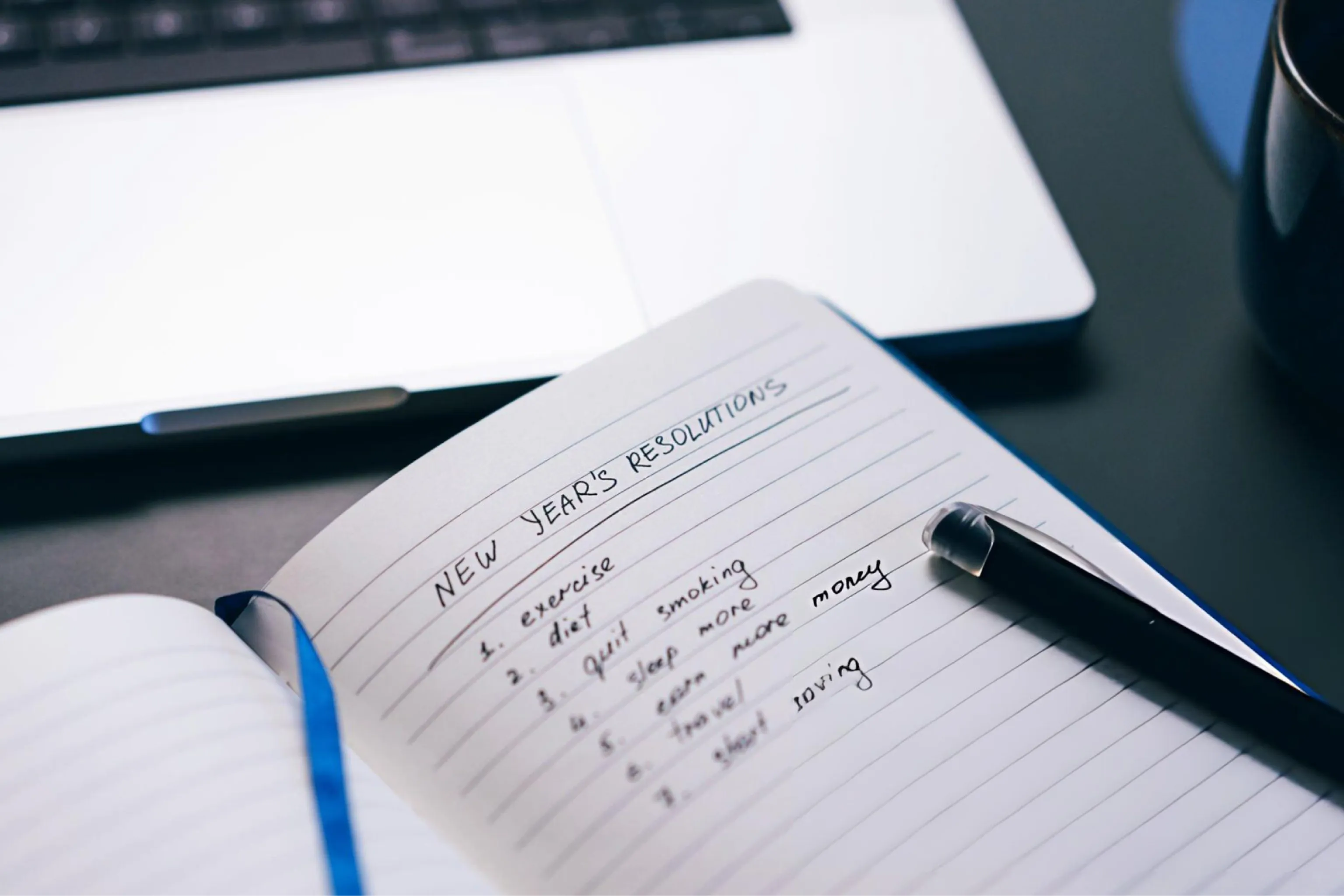 Open notebook lists “New Year’s Resolutions” beside a black pen, with a laptop and blue pencil nearby, creating a focused planning mood.