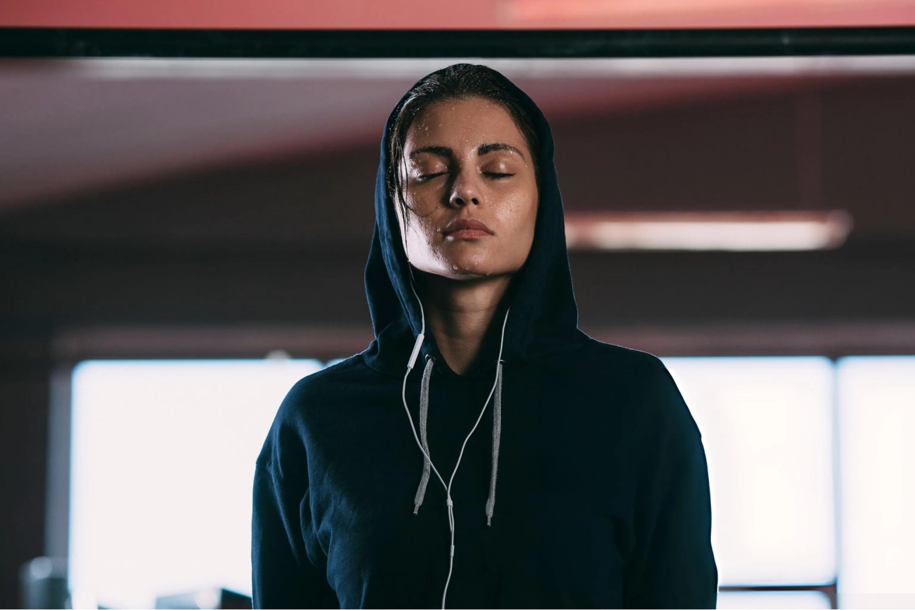 Young woman in a black hoodie stands with eyes closed and earbuds in a dim, modern space, conveying calm, focus, and introspection.