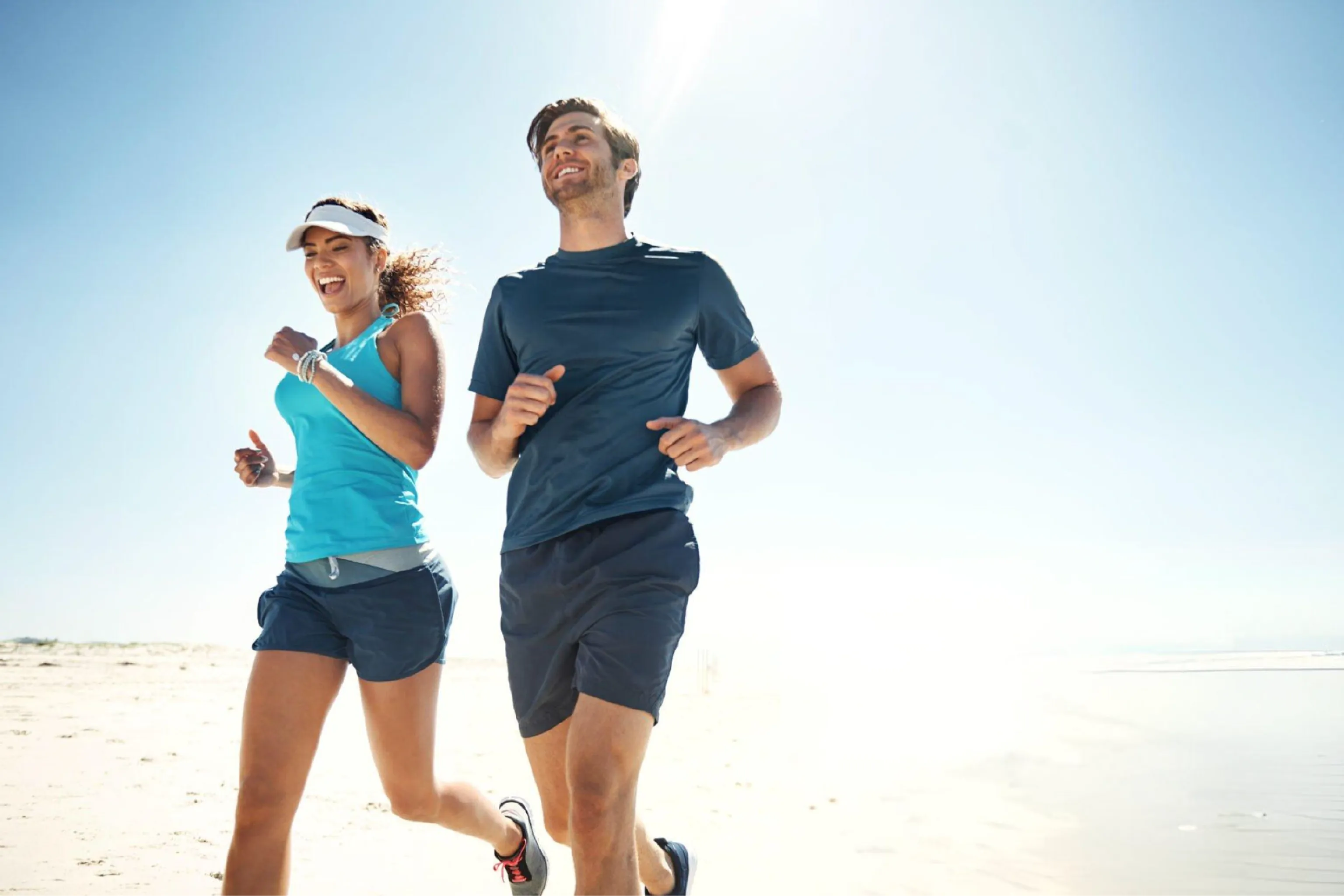 Smiling man and woman jog together along a sunlit beach in athletic clothes, with blue sky and ocean creating an energetic, joyful mood.