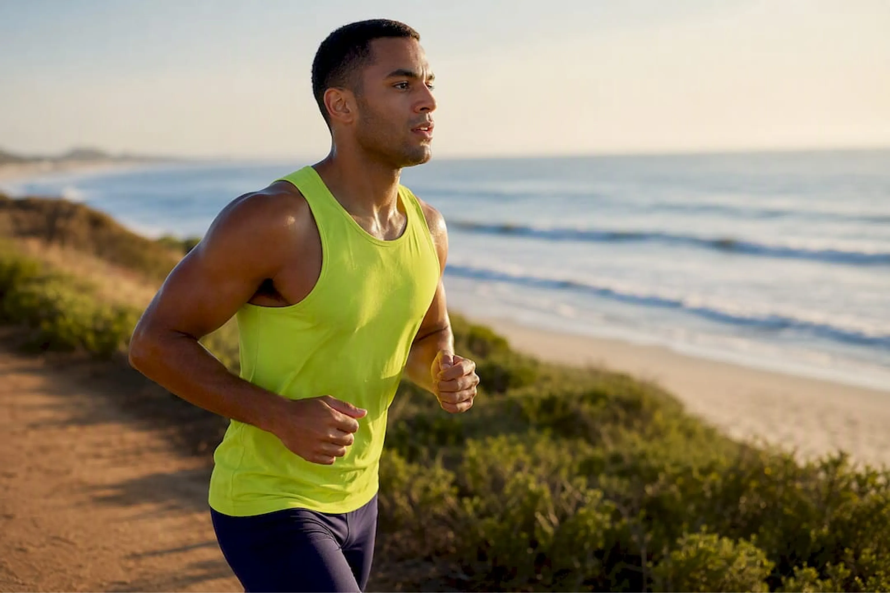 Athletic man in a neon yellow tank top jogs along a coastal trail beside a sandy beach and ocean, conveying energy, focus, and freedom.