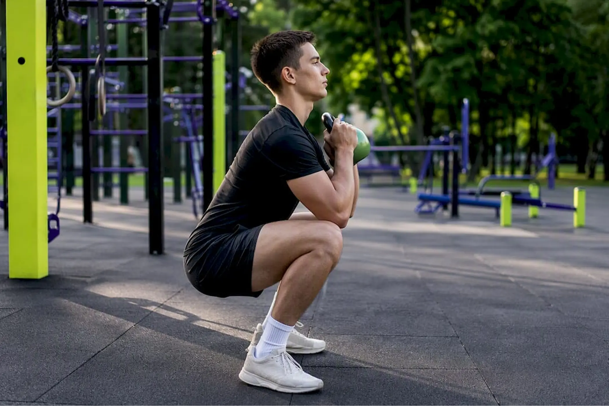 Man in black athletic wear performs a deep goblet squat with a kettlebell in an outdoor fitness park, surrounded by green and purple equipment.
