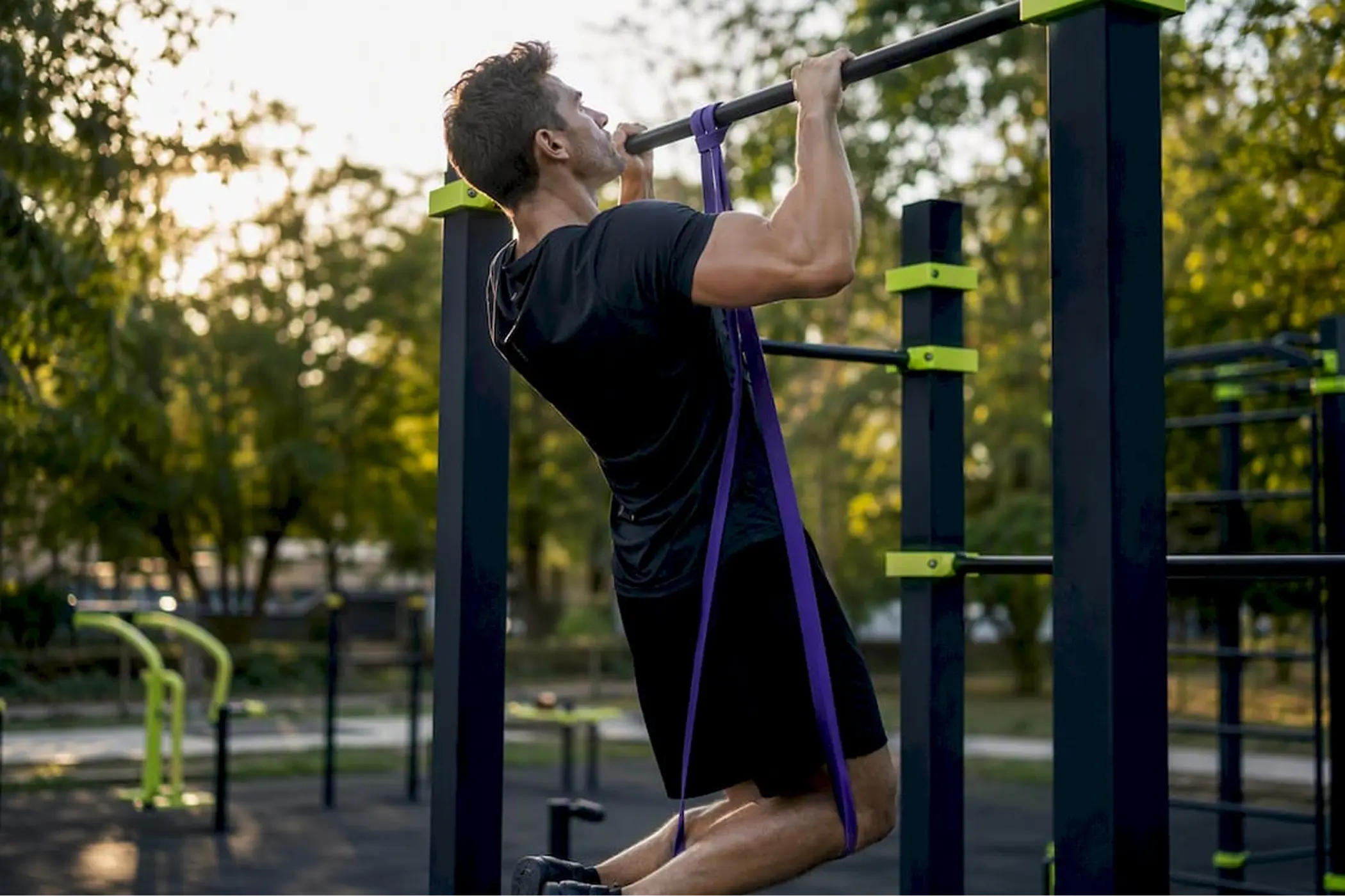 A man in dark athletic clothing performs a pull-up on outdoor fitness bars in a sunny park, surrounded by trees; the scene feels active and focused.