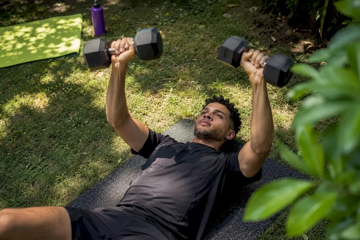 A bearded man in a black shirt lies on grass among leafy plants, lifting two weights overhead in a playful, energetic outdoor workout.