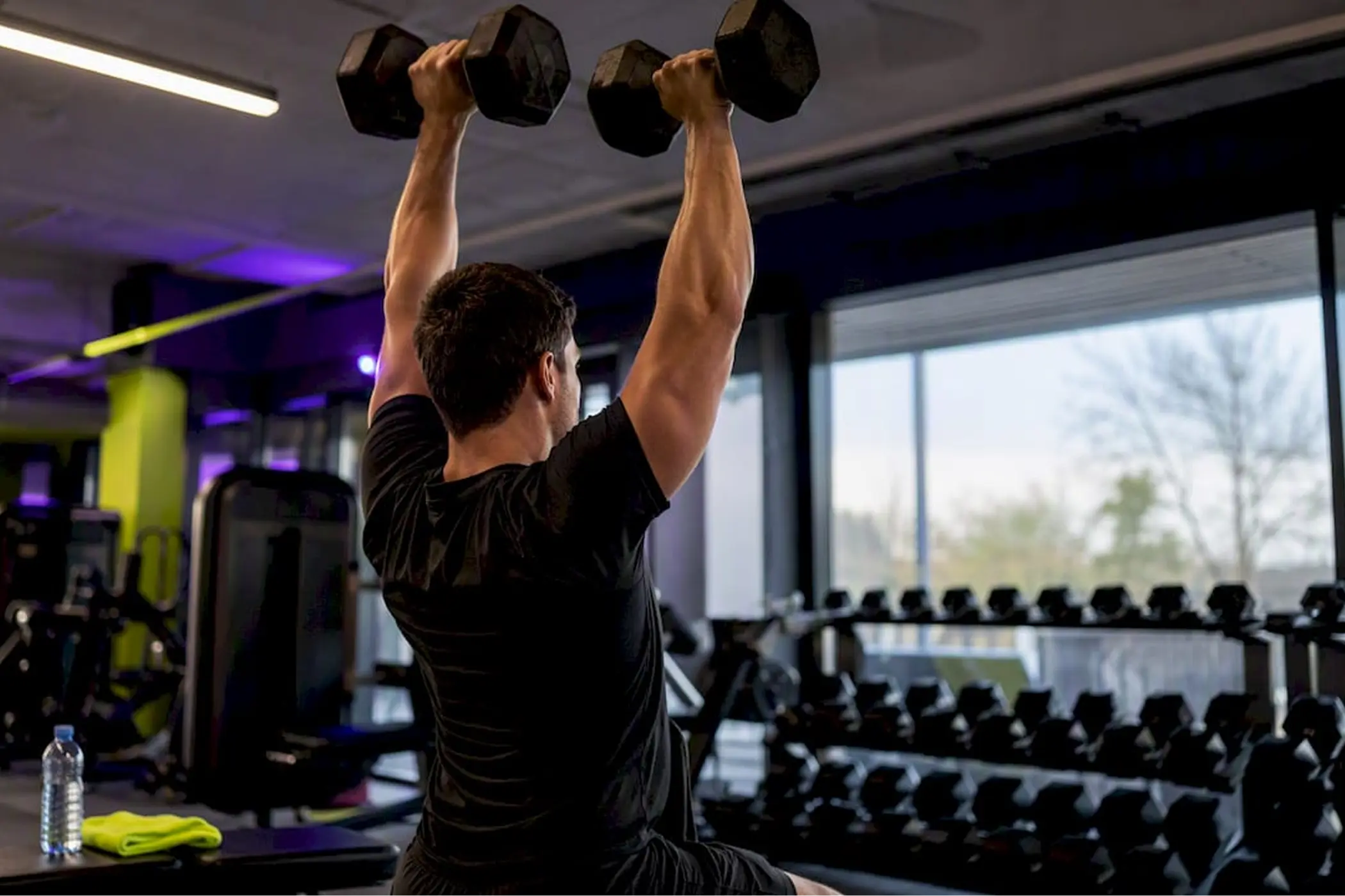 A focused man lifts two dumbbells overhead in a modern gym, with weight racks, large windows, and dramatic purple lighting behind him.