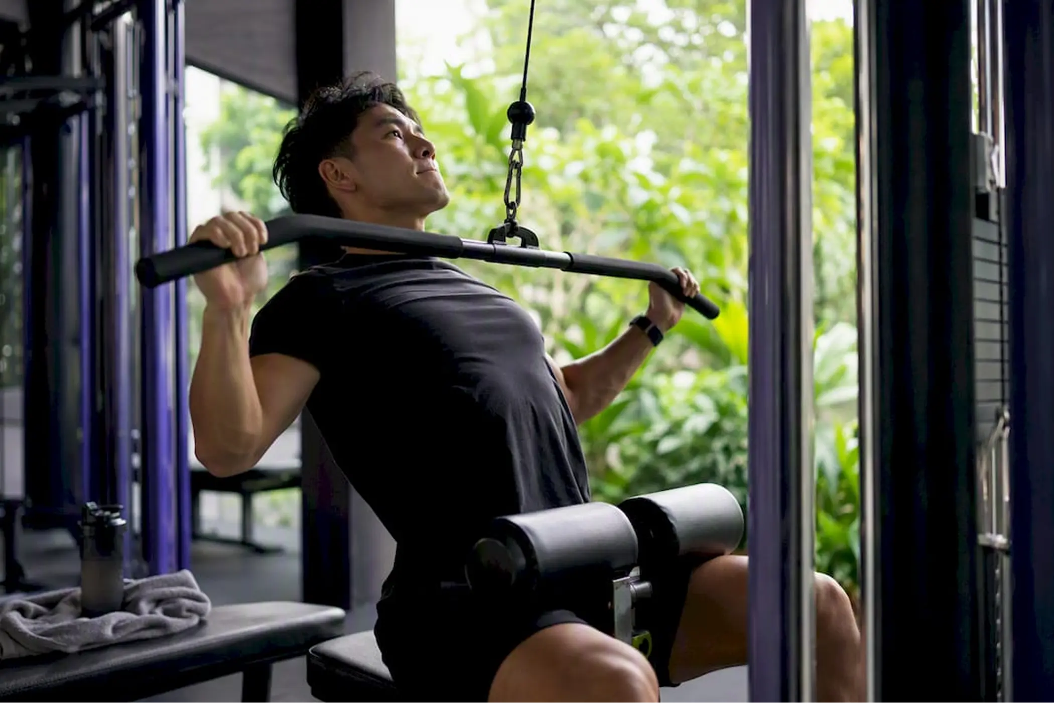 Man performs a seated cable-row exercise in a bright gym, gripping a bar with focused effort as green trees show through the window.