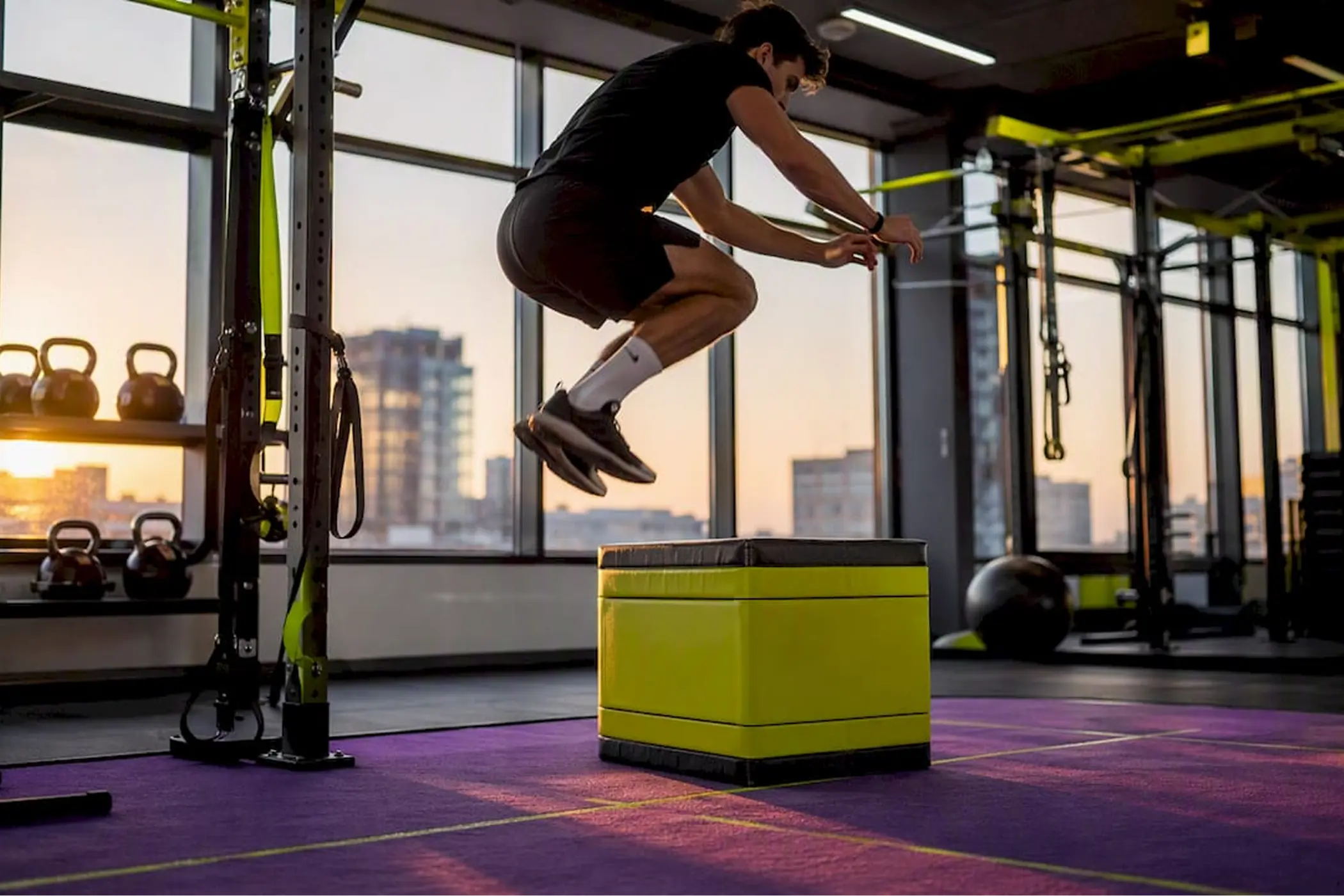Athlete in black workout clothes jumps onto a lime-green plyometric box in a modern gym, with kettlebells and city windows behind.