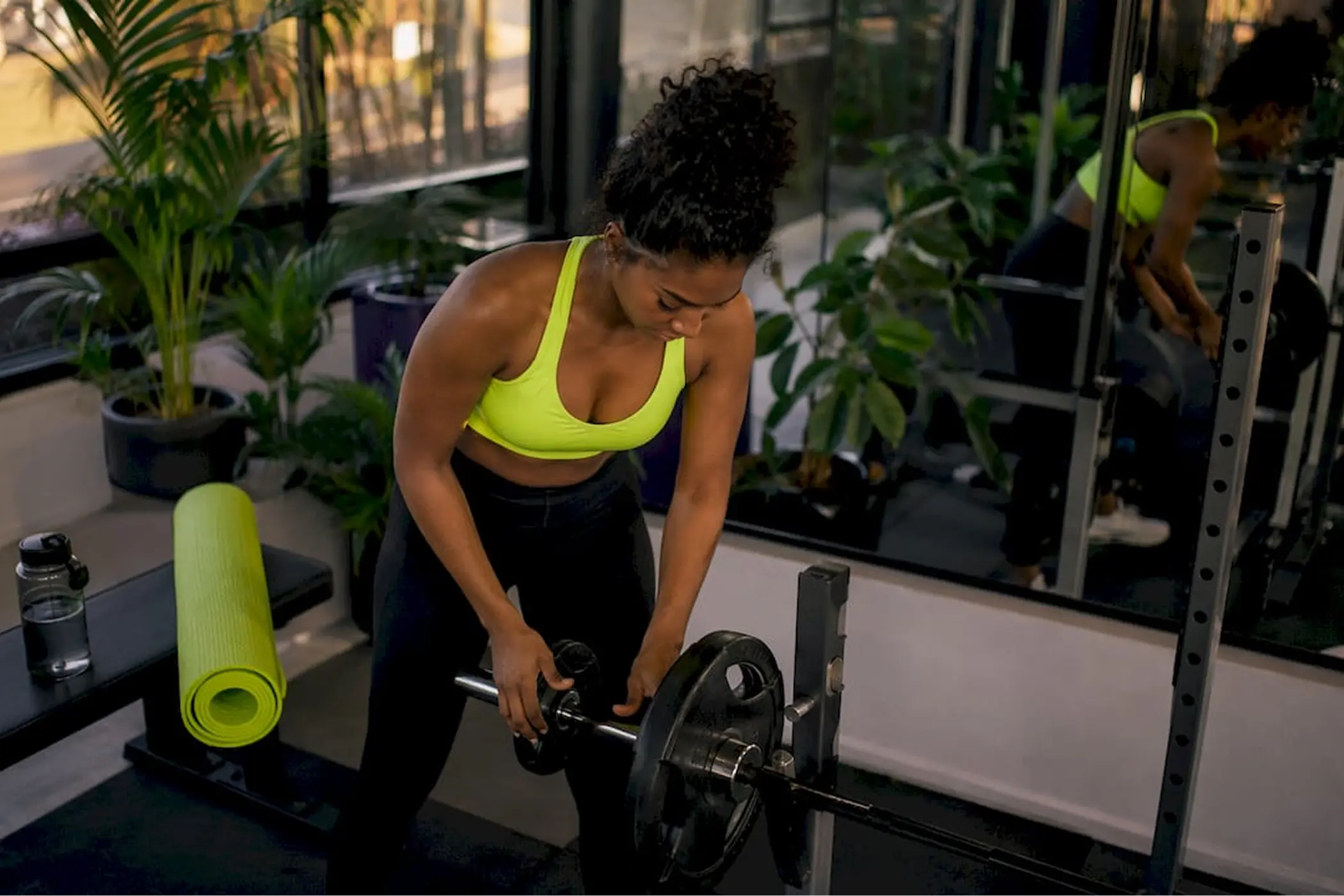 Focused woman in a neon green workout top loads a barbell in a bright, plant-filled gym, with mirrors and a rolled yoga mat nearby.