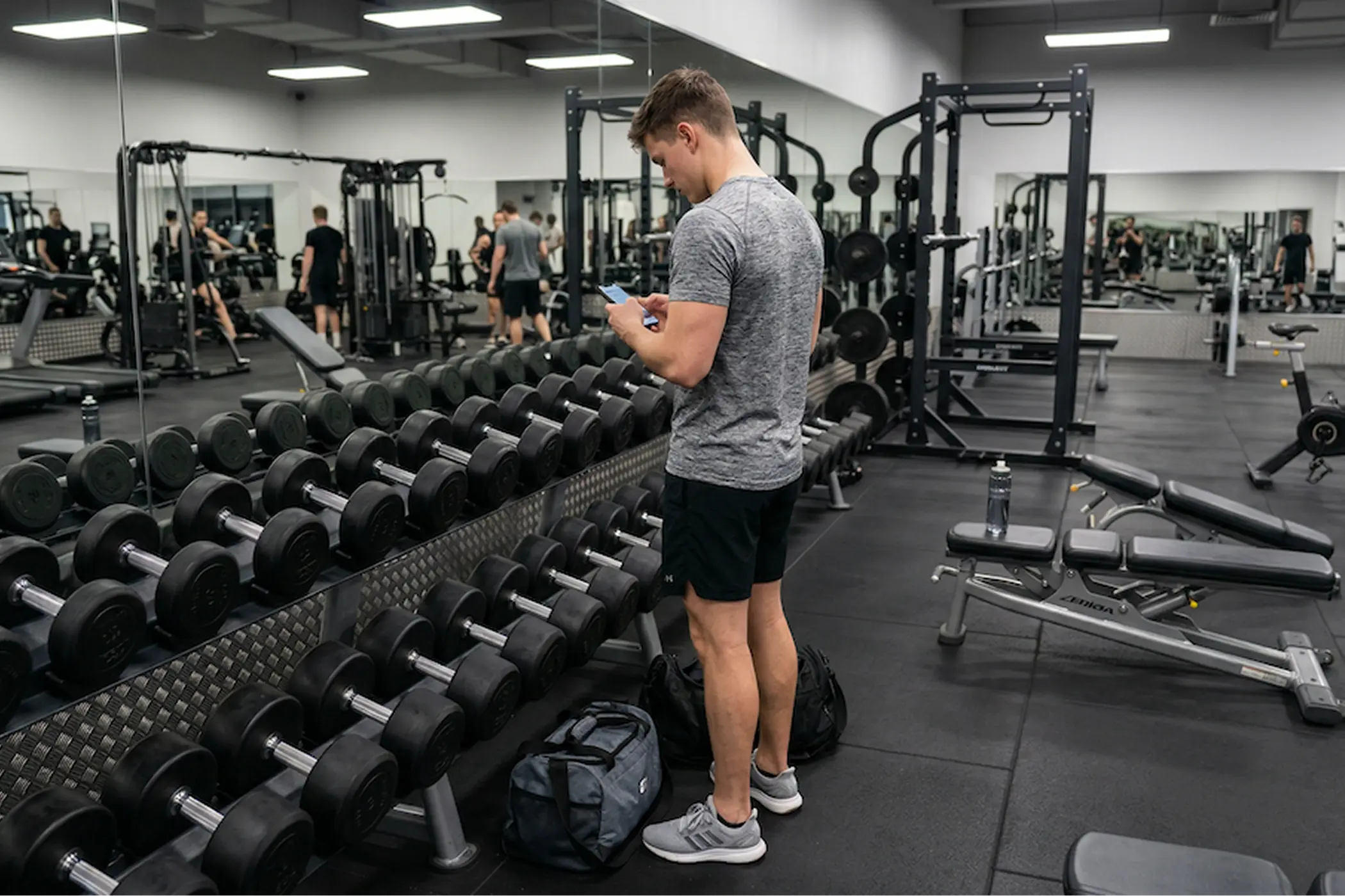 Man in a gray shirt checks his phone beside a dumbbell rack in a busy, well-equipped gym, creating a focused workout atmosphere.
