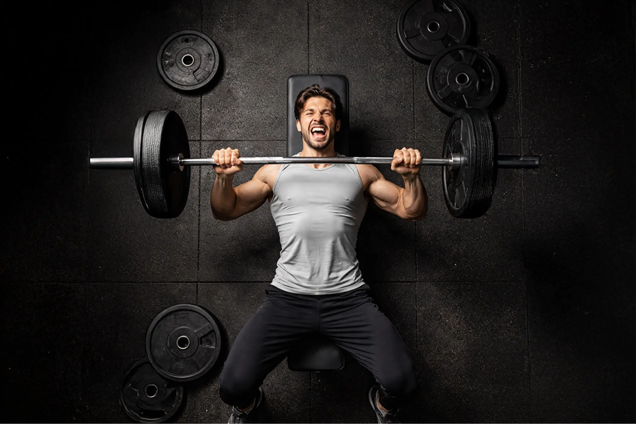 Muscular man in a gray shirt and black pants lifts a heavily weighted barbell, grinning with effort against a dark gym wall.