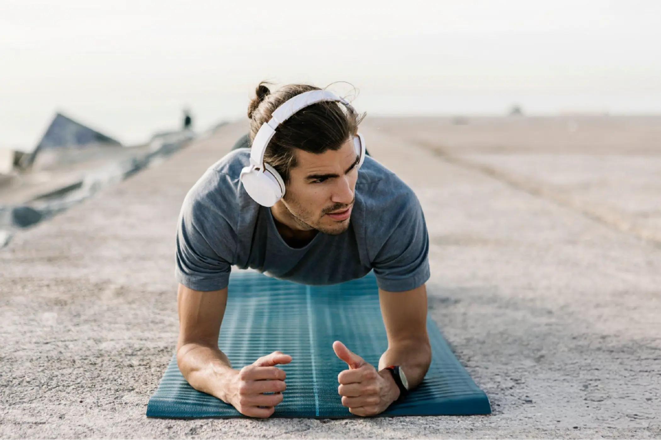 Man wearing white headphones holds a plank on a blue exercise mat outdoors, giving thumbs up with a focused, determined expression.