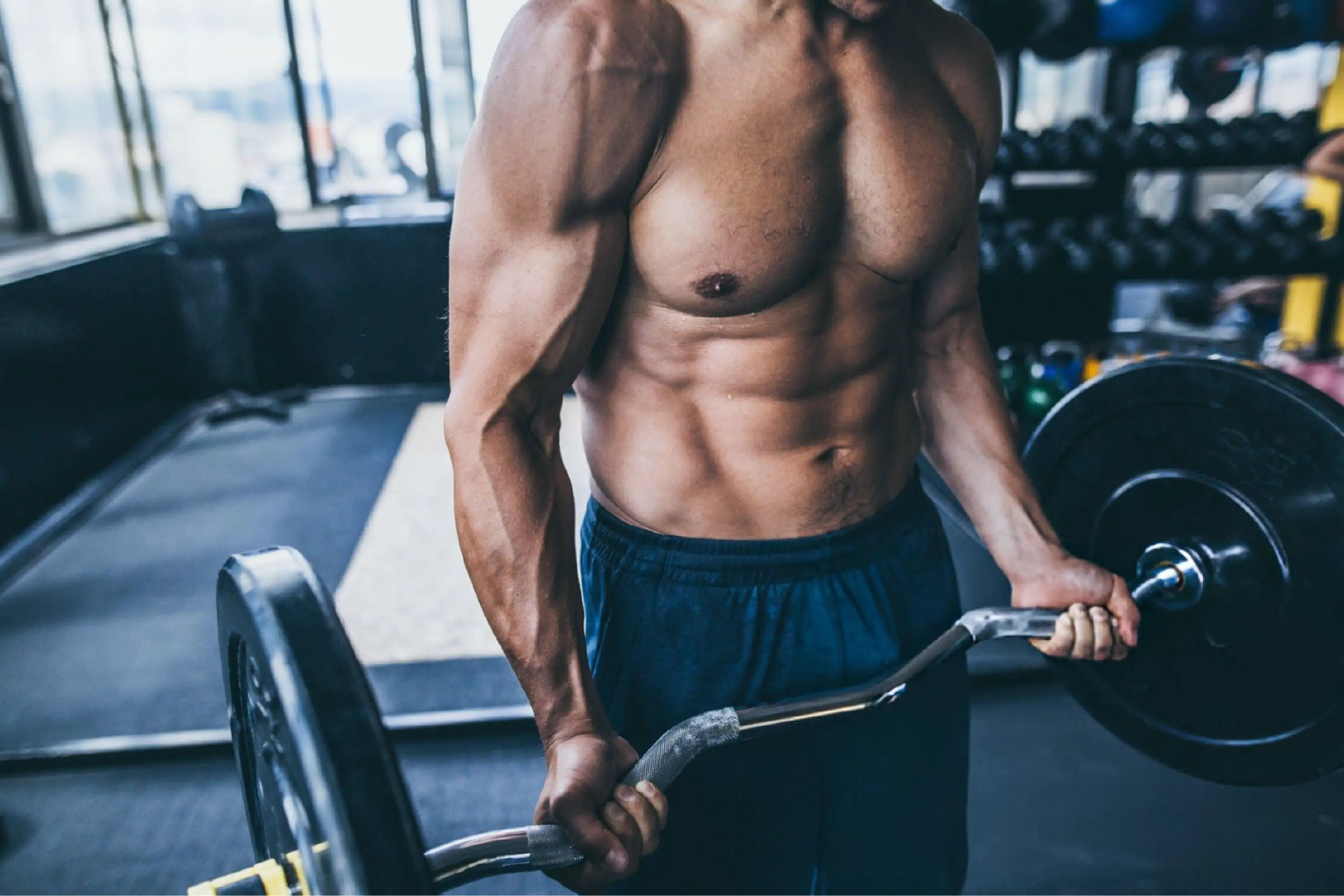 Muscular shirtless man performs a barbell curl in a gym, gripping the weight with both hands; dramatic light emphasizes strength and focus.
