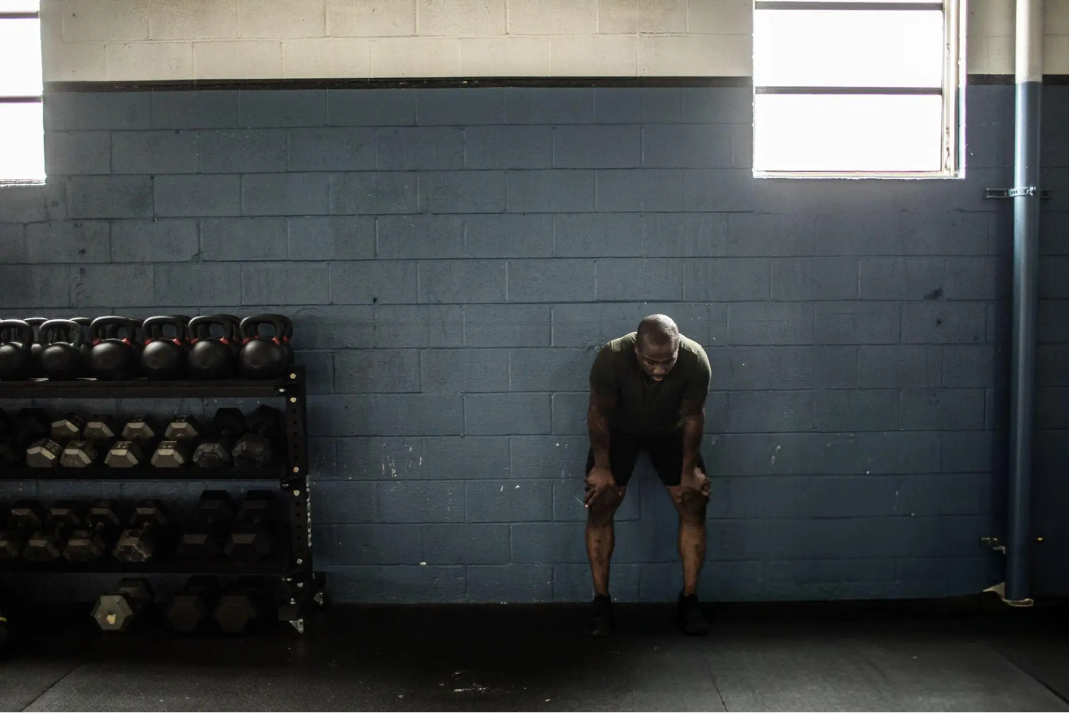 Exhausted athlete rests against a blue gym wall beside racks of kettlebells and dumbbells, with sunlight streaming through high windows.