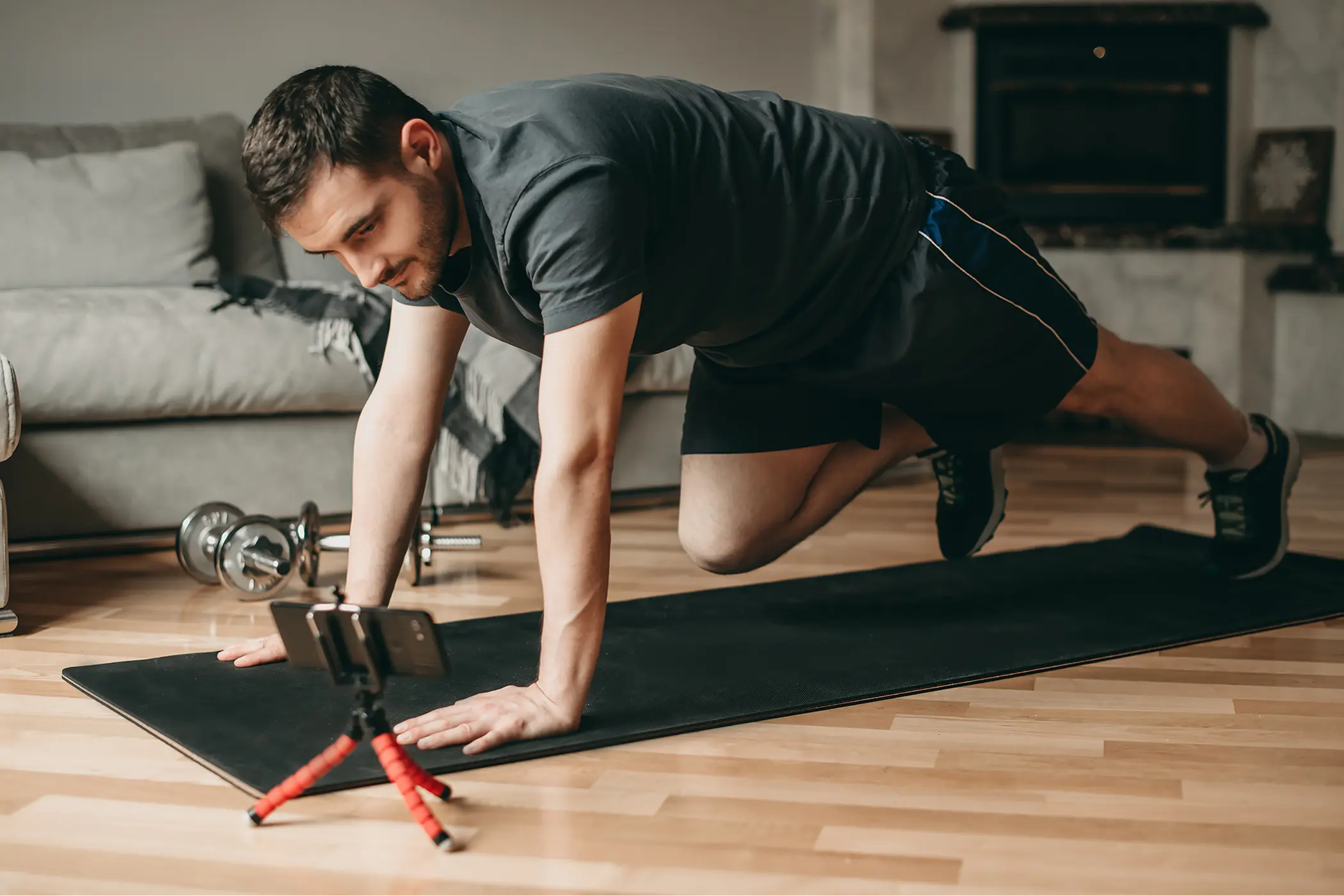 Man performs mountain climbers on a yoga mat at home, watching a smartphone on a mini tripod; dumbbells nearby create a focused workout scene.