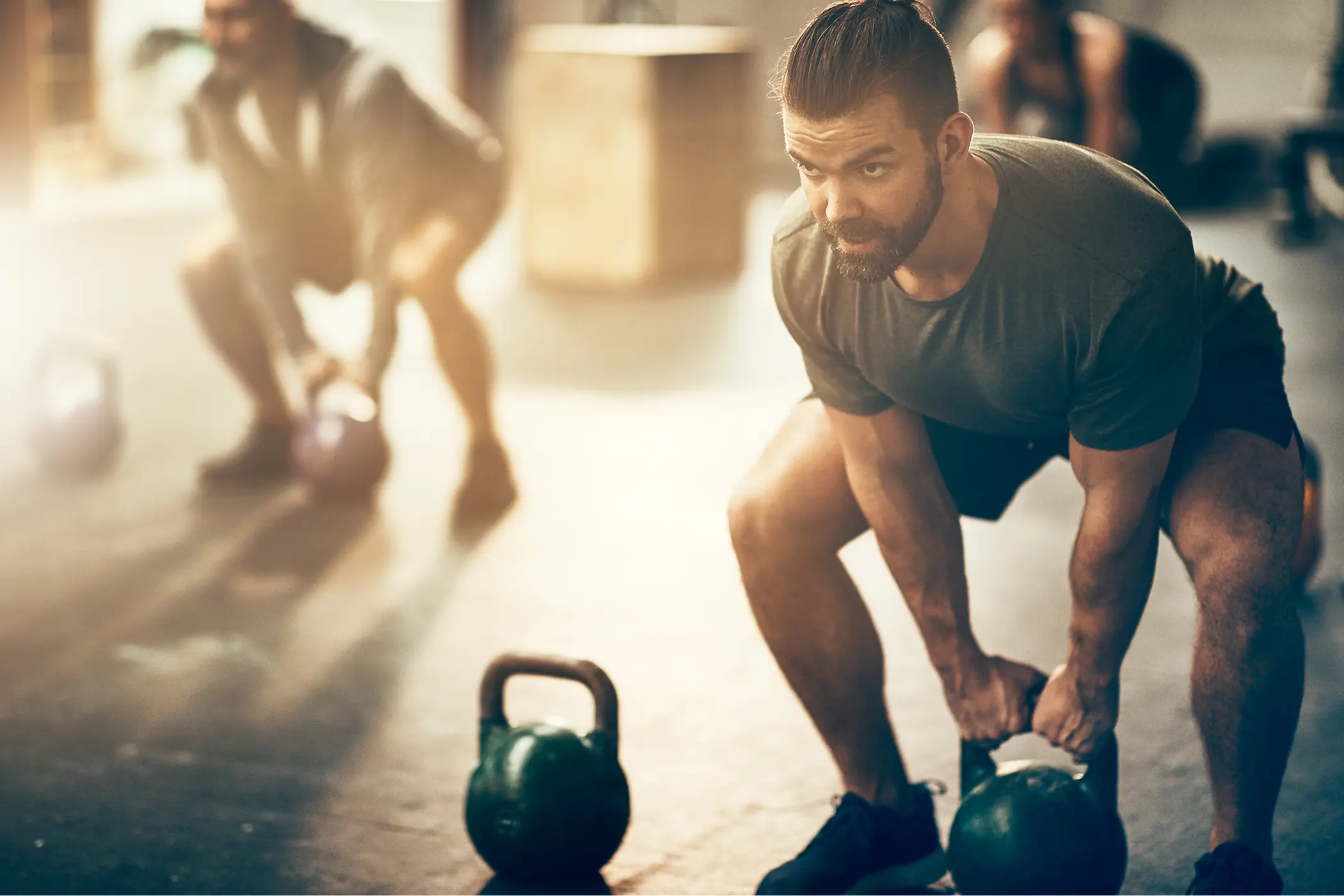 Bearded man lifting a kettlebell during an intense gym workout, with another athlete exercising in the sunlit background.