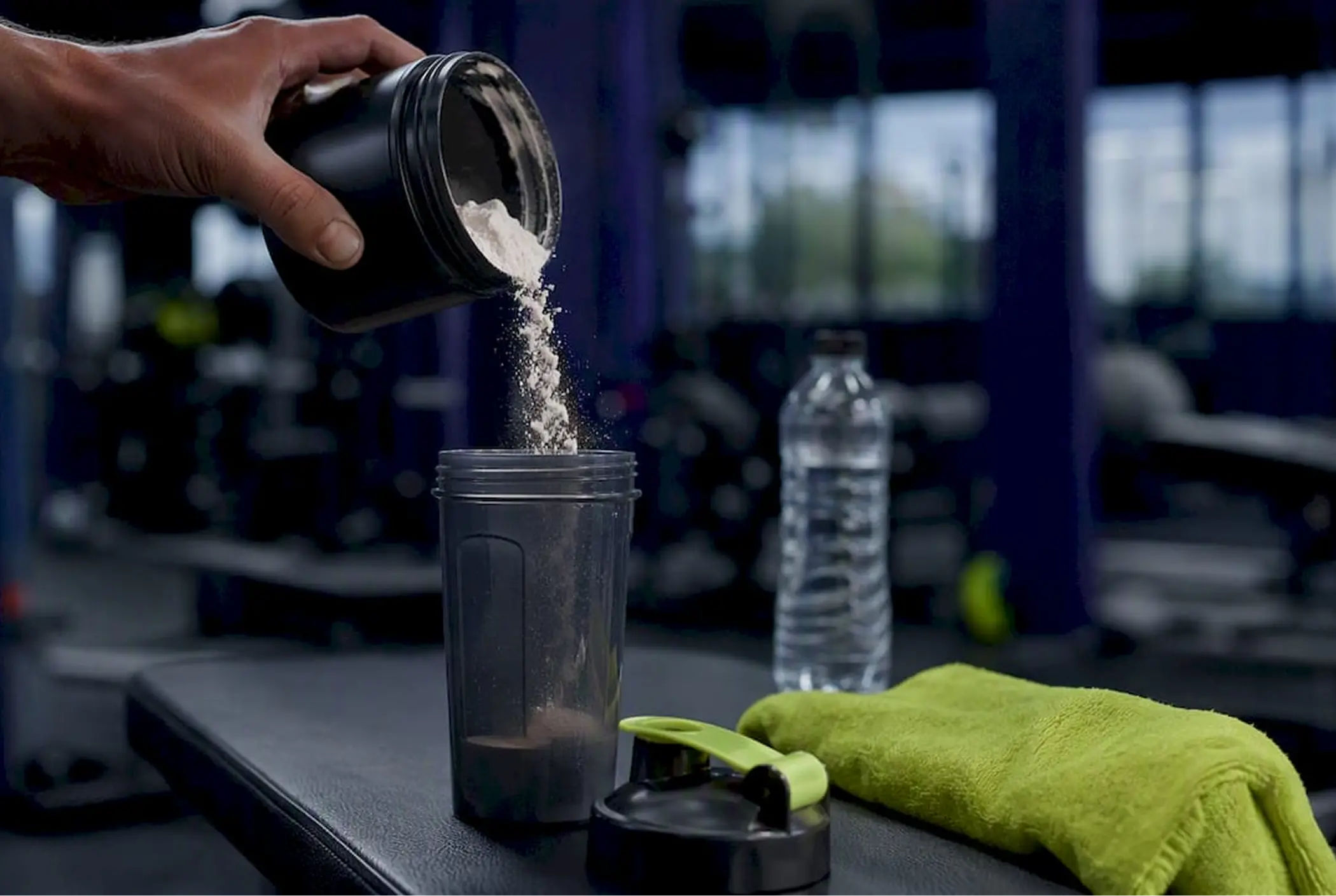Hand pours protein powder into a shaker bottle at a gym, beside a water bottle and green towel, creating a focused fitness scene.