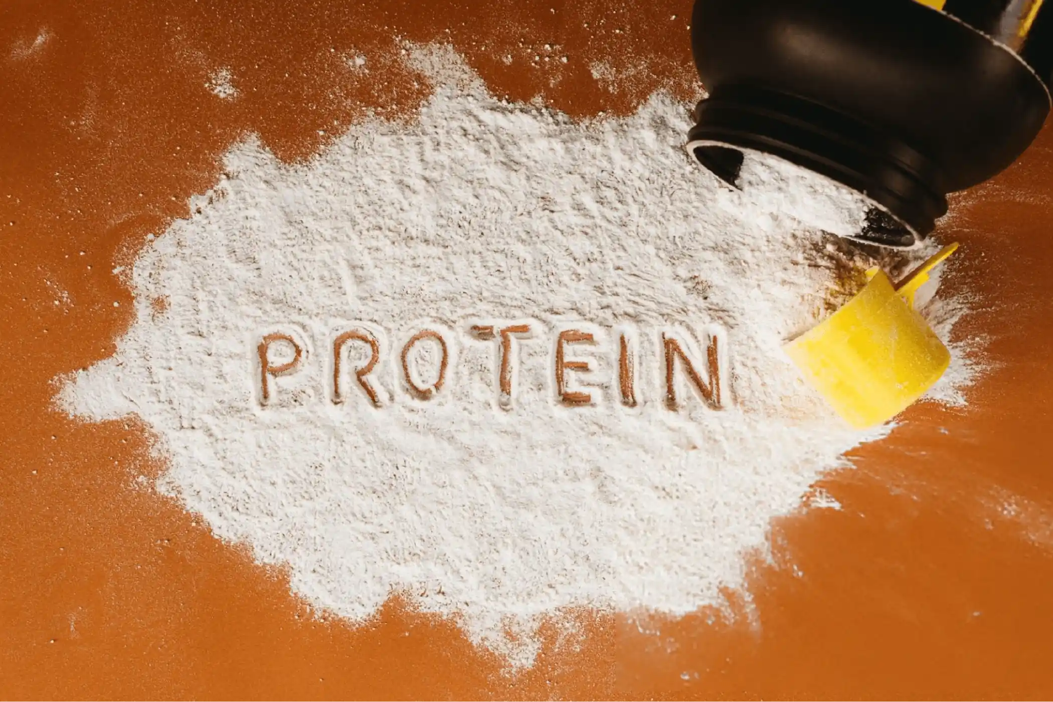 White protein powder forms a mound on a brown surface, with “PROTEIN” traced in it and a black-and-yellow scoop spilling powder.