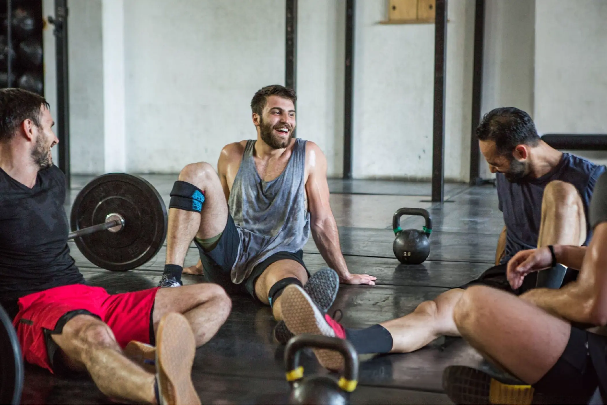 Four exhausted men sit on a gym floor after training, surrounded by barbells and kettlebells; one laughs, creating a friendly, energetic mood.