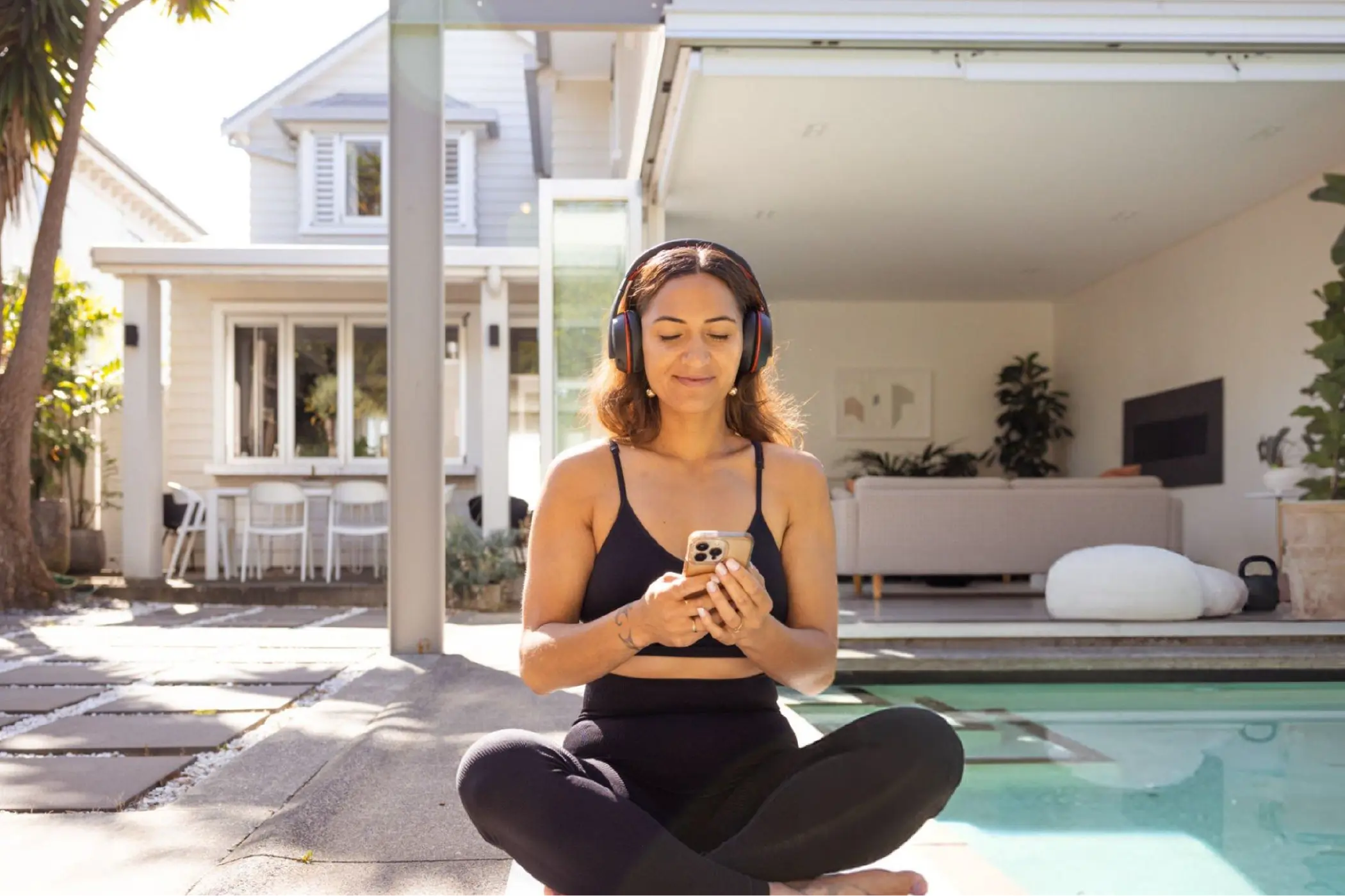 Woman in headphones sits cross-legged by a swimming pool, using her phone outside a modern home; bright sunlight creates a calm mood.