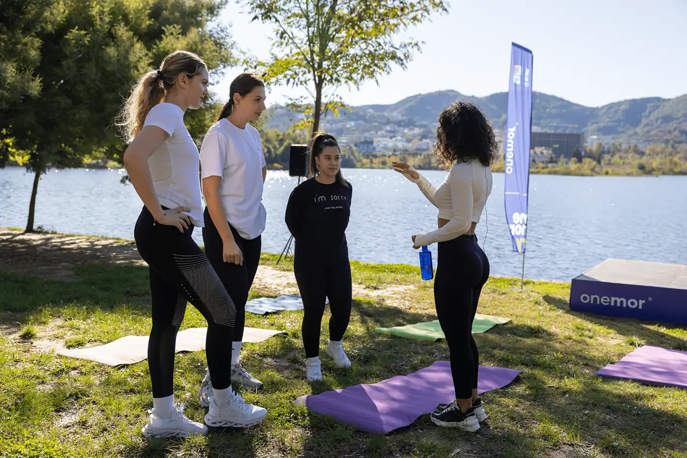 Four women gather for a friendly lakeside fitness class, chatting beside yoga mats, with trees, mountains, water, and an Onemor banner.