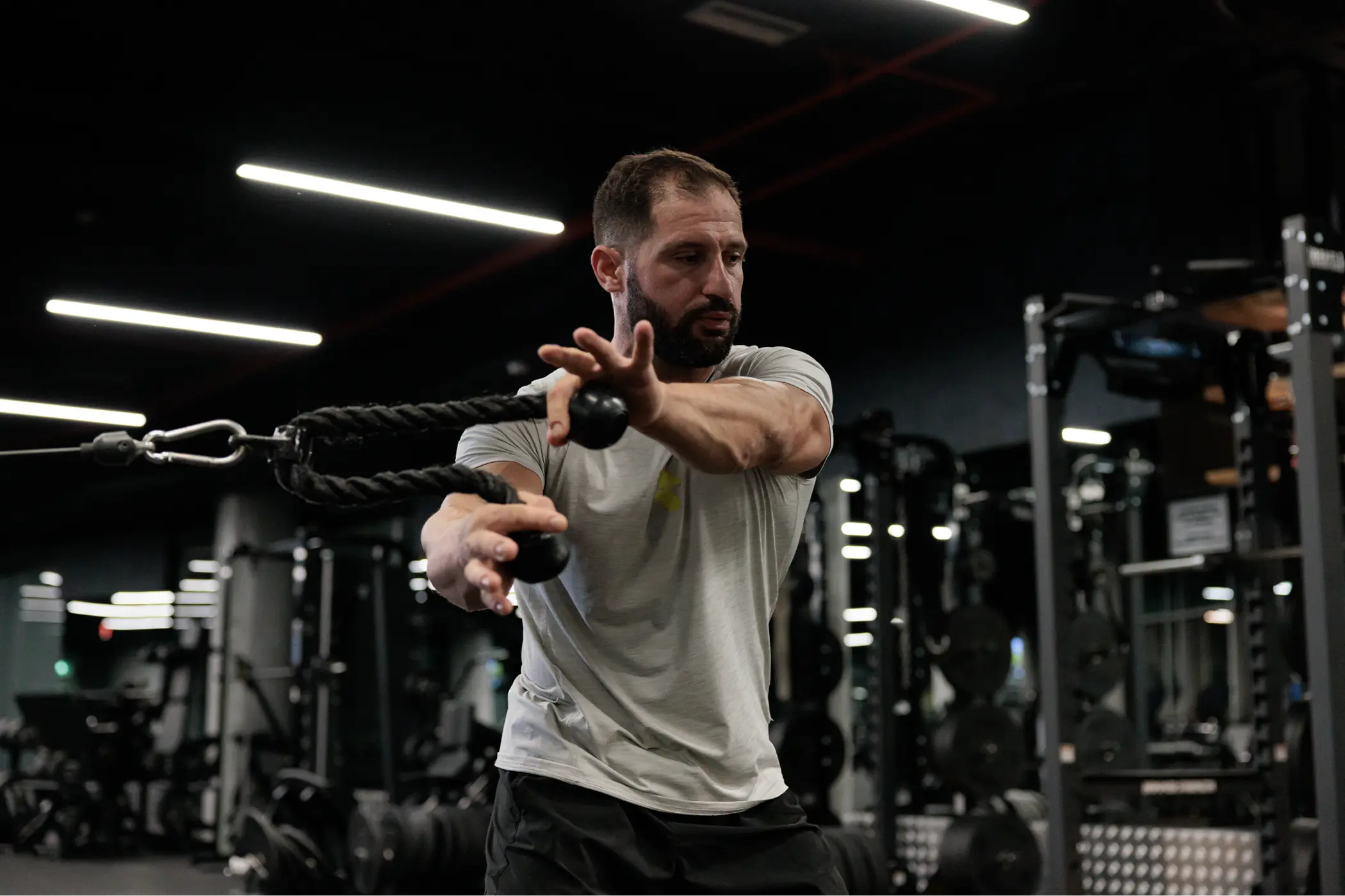 Bearded man performs a cable-rope exercise in a dimly lit gym, reaching forward with focus as weight machines fill the background.