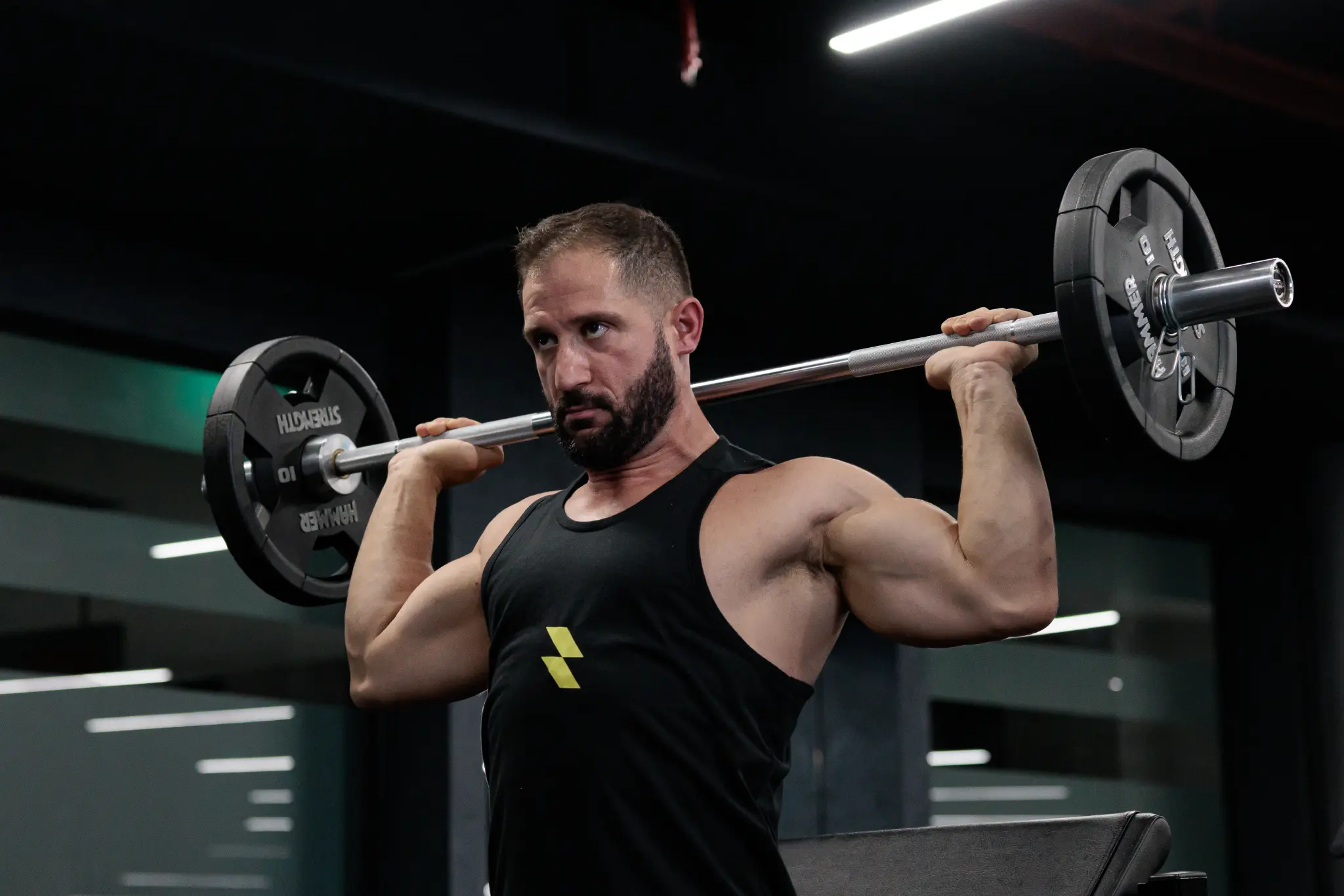 Muscular bearded man in a black tank top braces a loaded barbell across his shoulders in a dim gym, conveying focus and strength.
