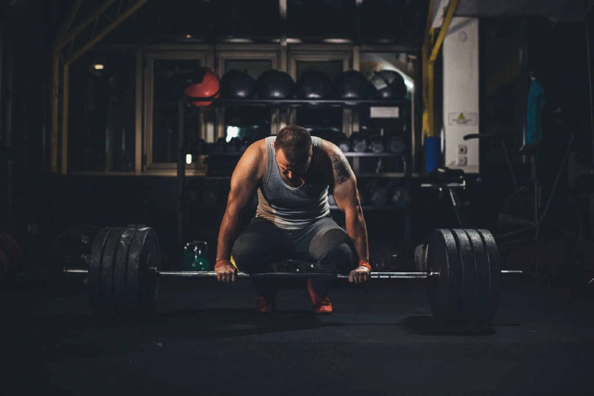 Muscular athlete crouches over a heavily loaded barbell in a dim gym, gripping it before a deadlift; dramatic lighting conveys focus and strain.