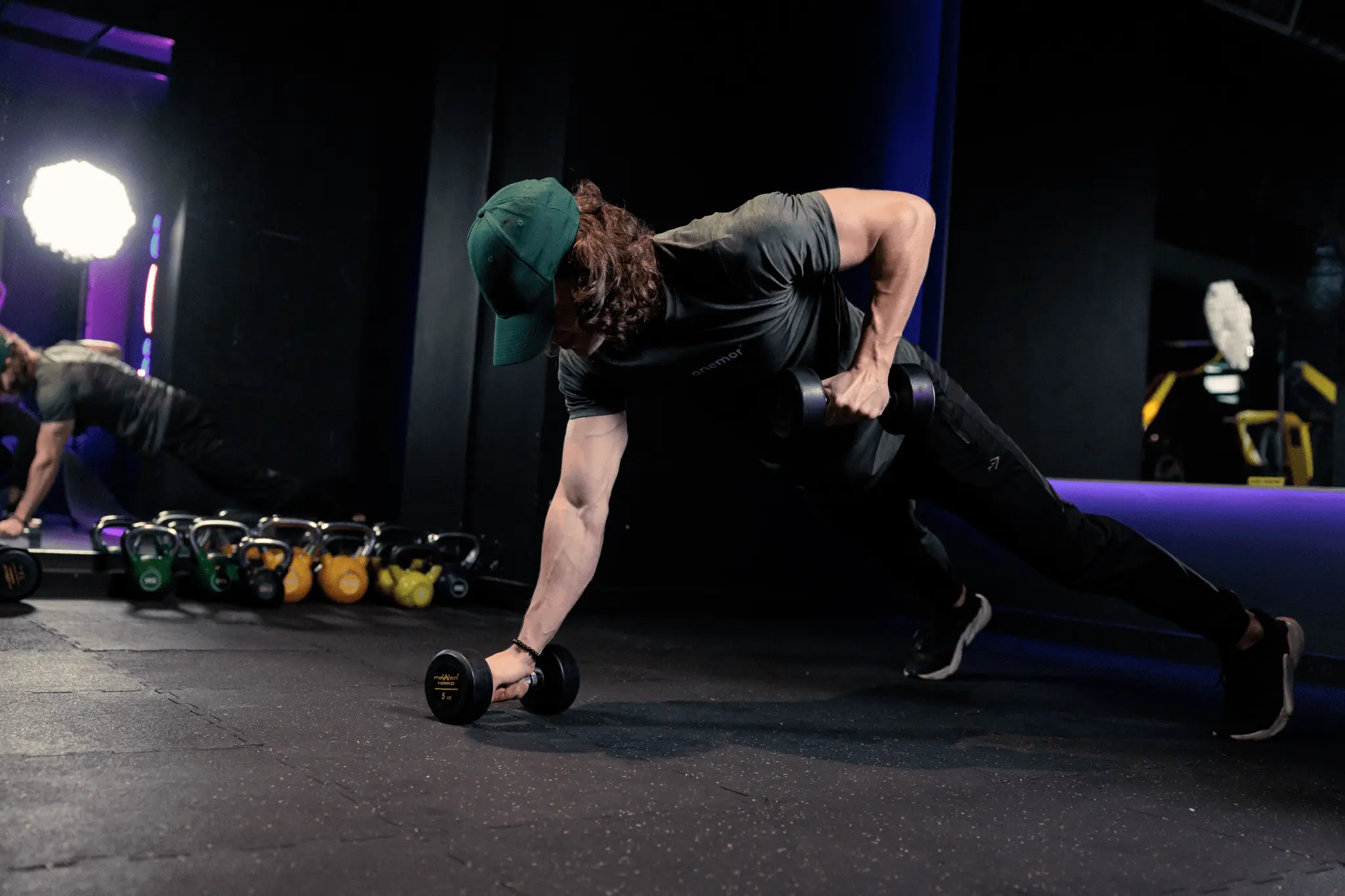 Muscular athlete performs a renegade row with dumbbells in a dark gym, surrounded by kettlebells and mirrors, conveying strength and focus.