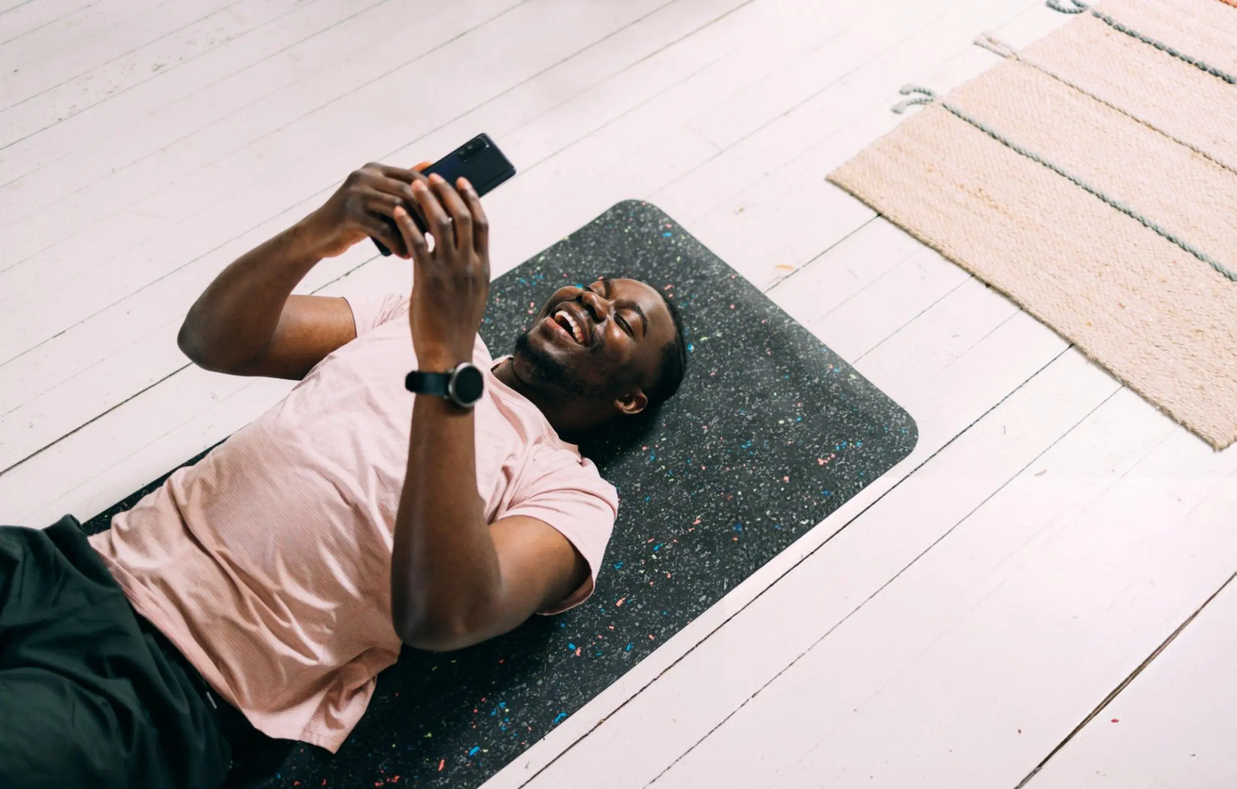 Smiling Black man in a pink shirt lies on a black exercise mat, holding a smartphone above him on a bright wood floor, relaxed and happy.