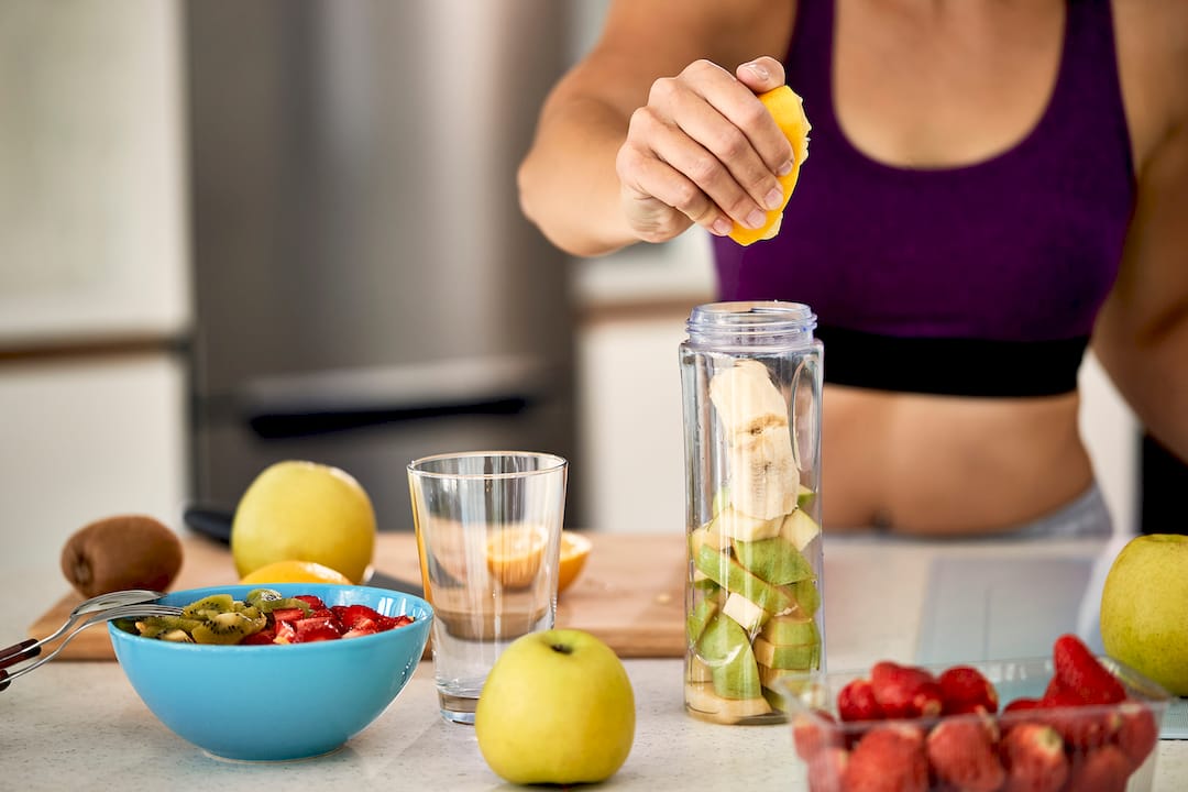 Woman in workout clothes squeezes lemon into a blender filled with fruit, surrounded by fresh produce, conveying a healthy lifestyle.