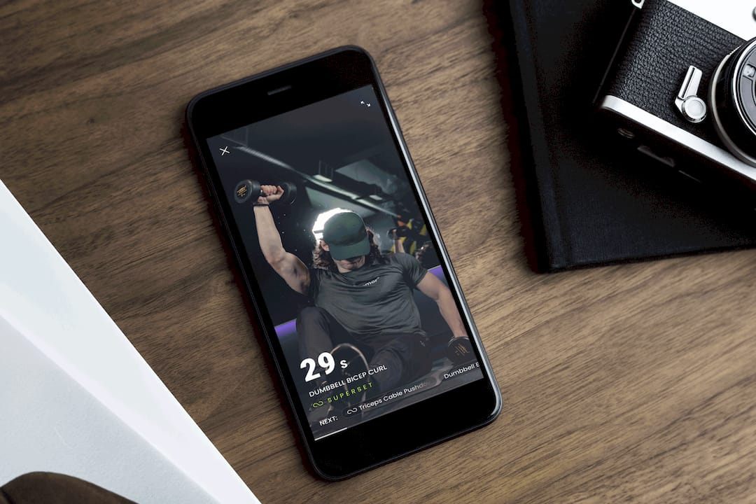 Smartphone on a wooden desk displays a fitness app workout with a helmeted athlete lifting weights; a camera and notebook sit nearby.