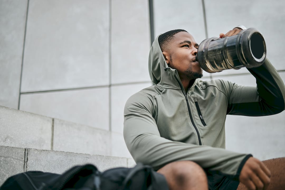 Athletic man in a gray hooded jacket drinks from a large water bottle outdoors, seated beside a backpack against a modern concrete wall.