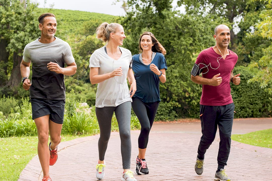 Four diverse adults jog together on a tree-lined park path, smiling and chatting; sunny outdoor scene conveys friendship, fitness, and energy.
