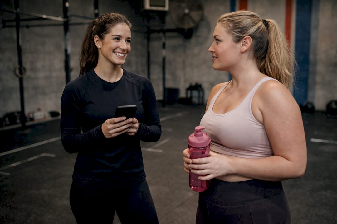 Two smiling women chat in an industrial gym: one holds a smartphone, while the other carries a pink water bottle after a workout.