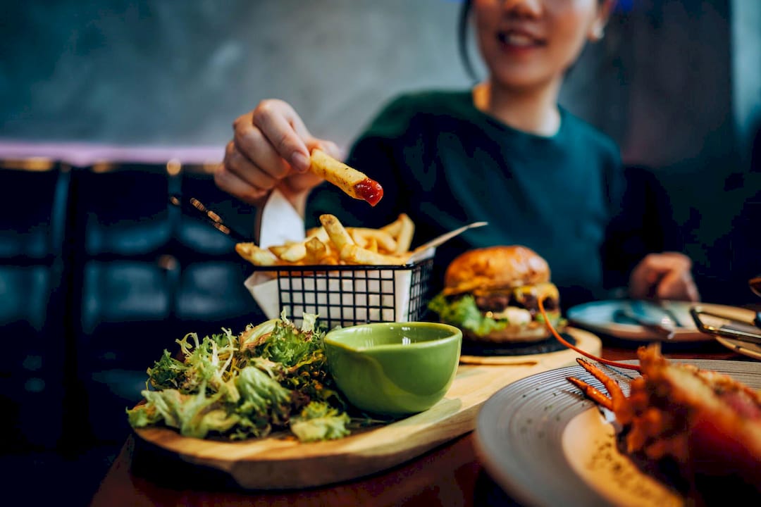 Smiling diner dips a golden fry into sauce beside a burger, salad, and green bowl in a warmly lit restaurant, creating a cheerful dining scene.