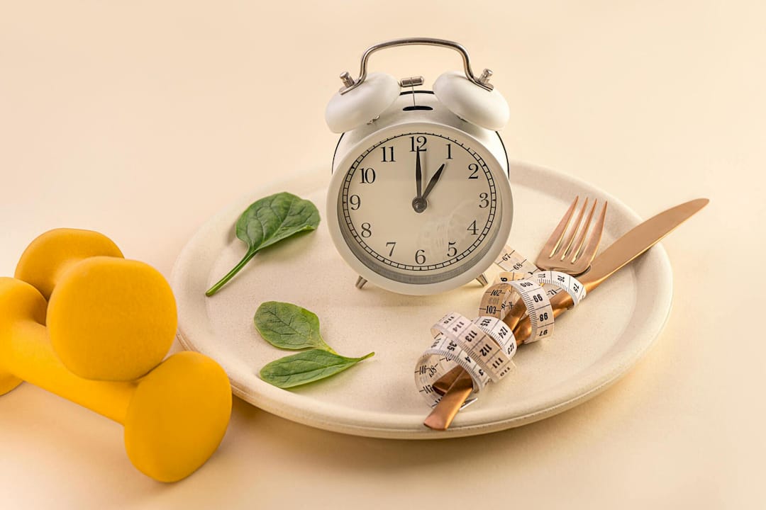 Still life of a vintage alarm clock on a plate with mango slices, spinach leaves, fork, knife, and measuring tape, suggesting timed dieting.