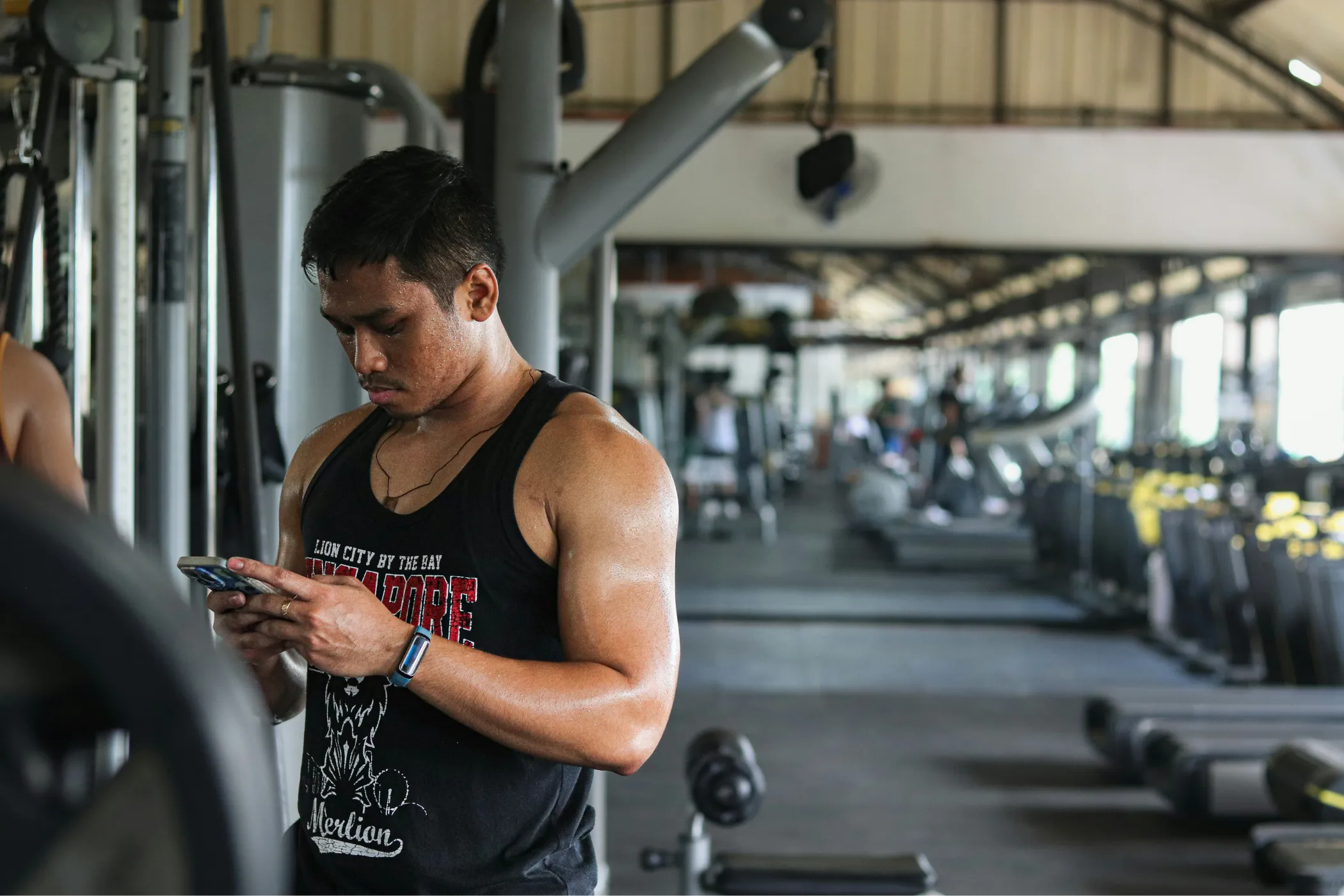 A focused man in a black tank top checks his phone beside weight machines in a busy gym, taking a quiet break amid the workout activity.