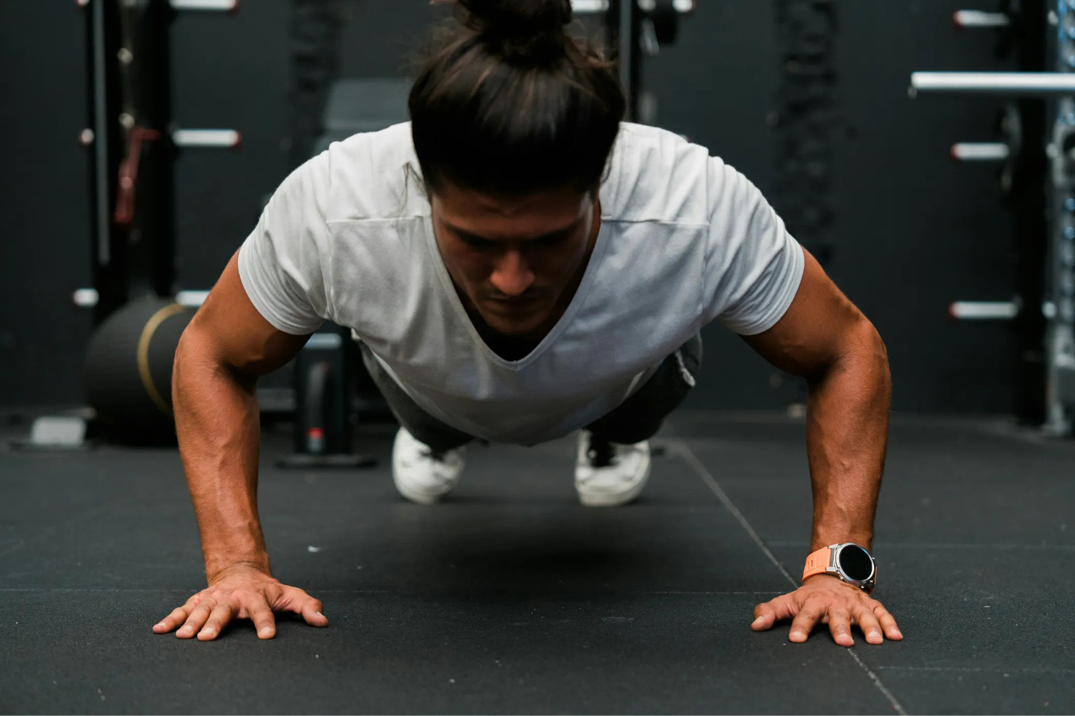 A focused athlete with hair in a bun holds a push-up position on a gym floor, hands planted wide as weight equipment fills the dark background.