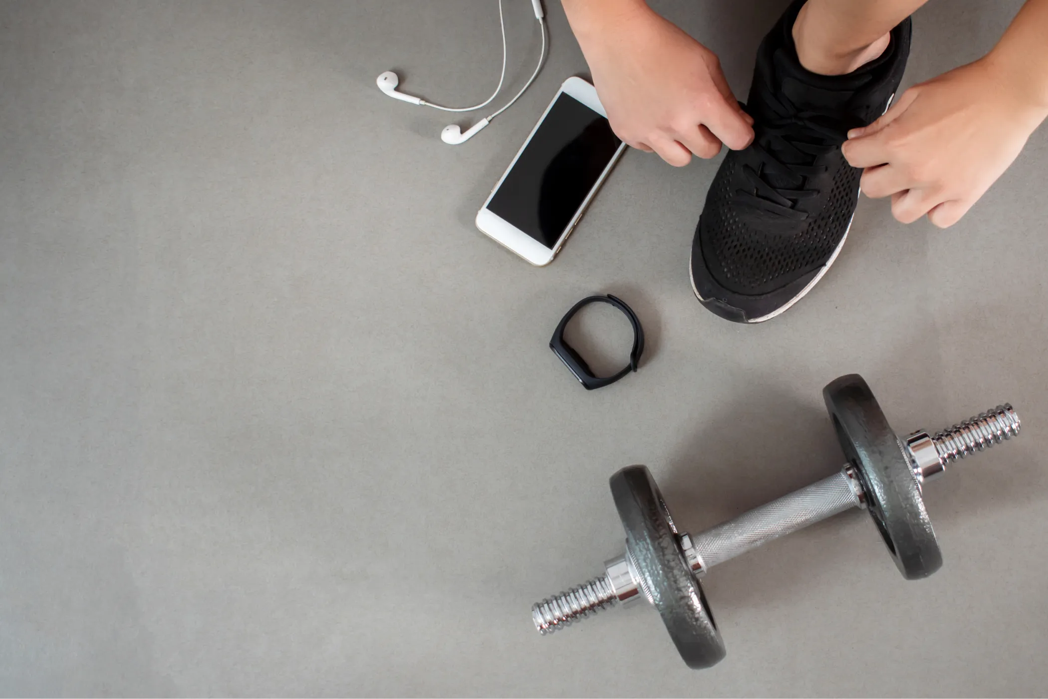 Top-down view of a person tightening a black running shoe beside a smartphone, earbuds, fitness tracker, and dumbbell, suggesting preparation for a workout.