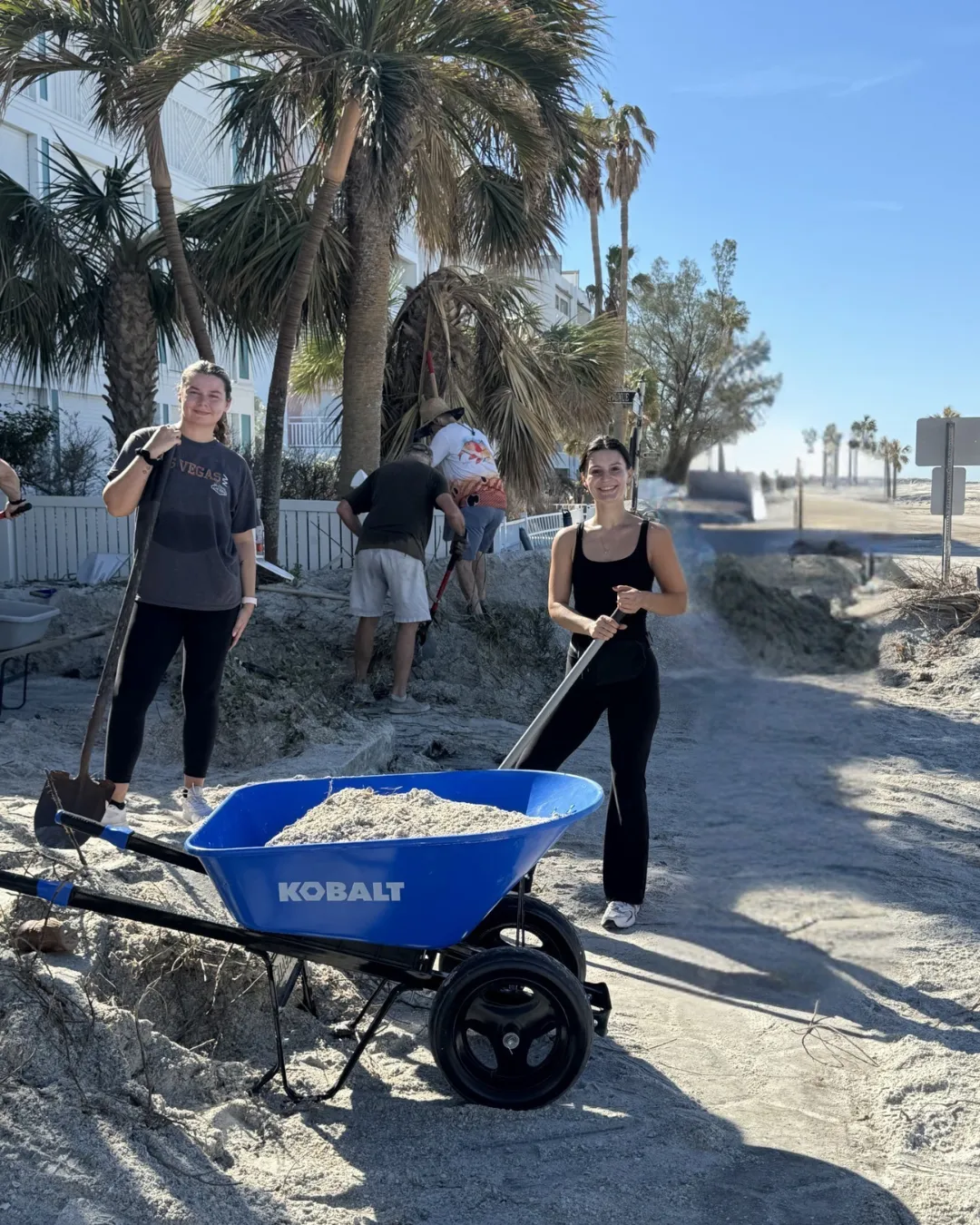 An image from our St. Pete Marketing Agency team beach clean, two young women hold shovels behind a wheel barrel of sand